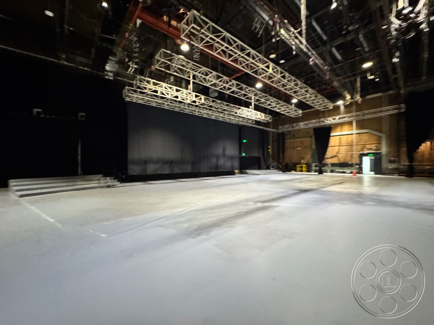 Stage 33 - concrete floor, exposed industrial ceiling, acoustic wall panels, interior soundstage, black drapery wall, metal truss ceiling, pendant stage lighting, gray non-slip studio flooring, elevated stage platform, dimly lit production space, studio control room access, open layout recording area, large rehearsal space, temporary backdrop setup