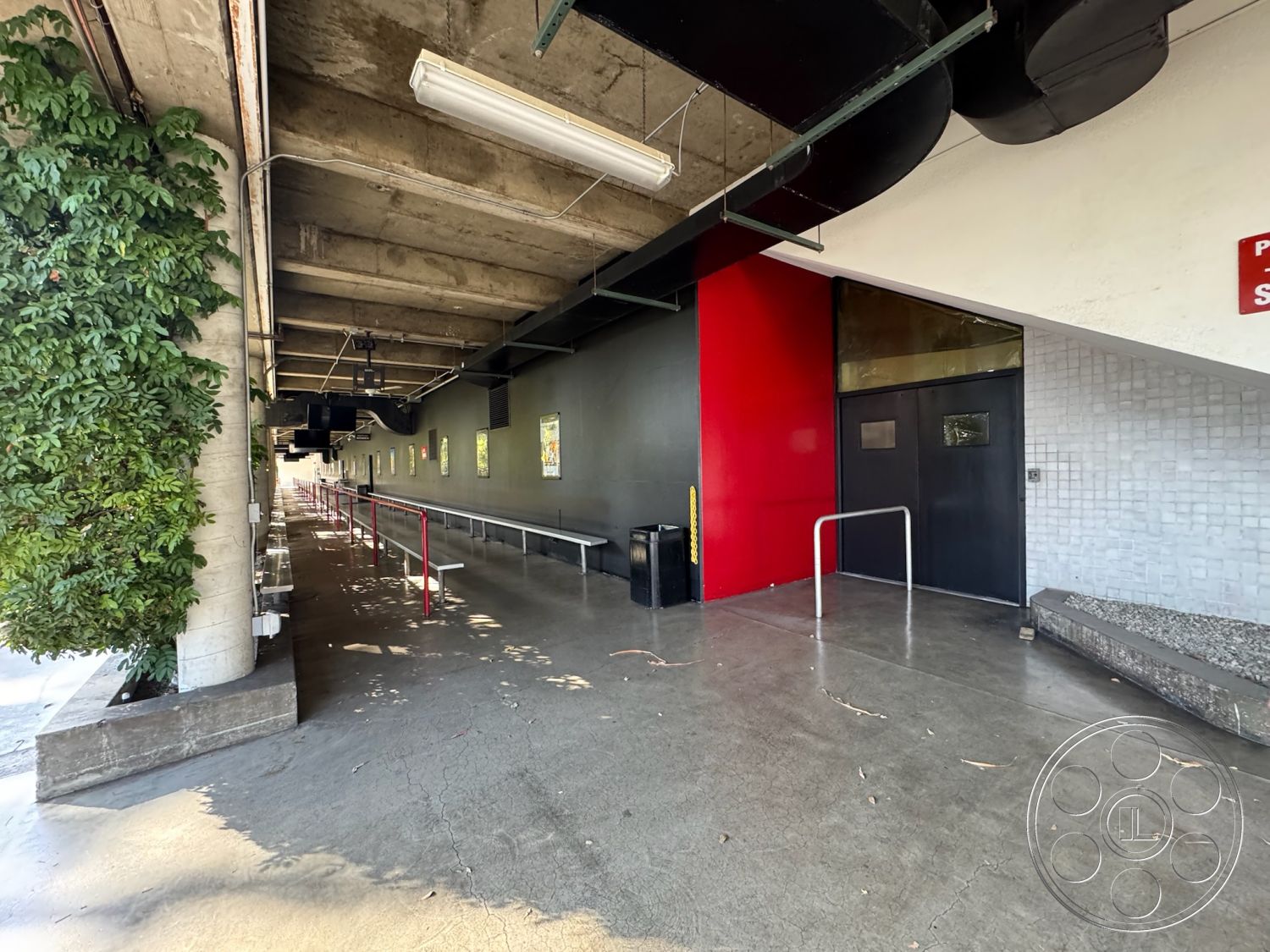 Stage 33 - concrete flooring, metal railing, landscaped greenery, urban setting, red accent wall, industrial design elements, modern architectural style, exposed concrete ceiling, exterior walkway, open-air corridor, black entrance door, glass door window, underground parking lot