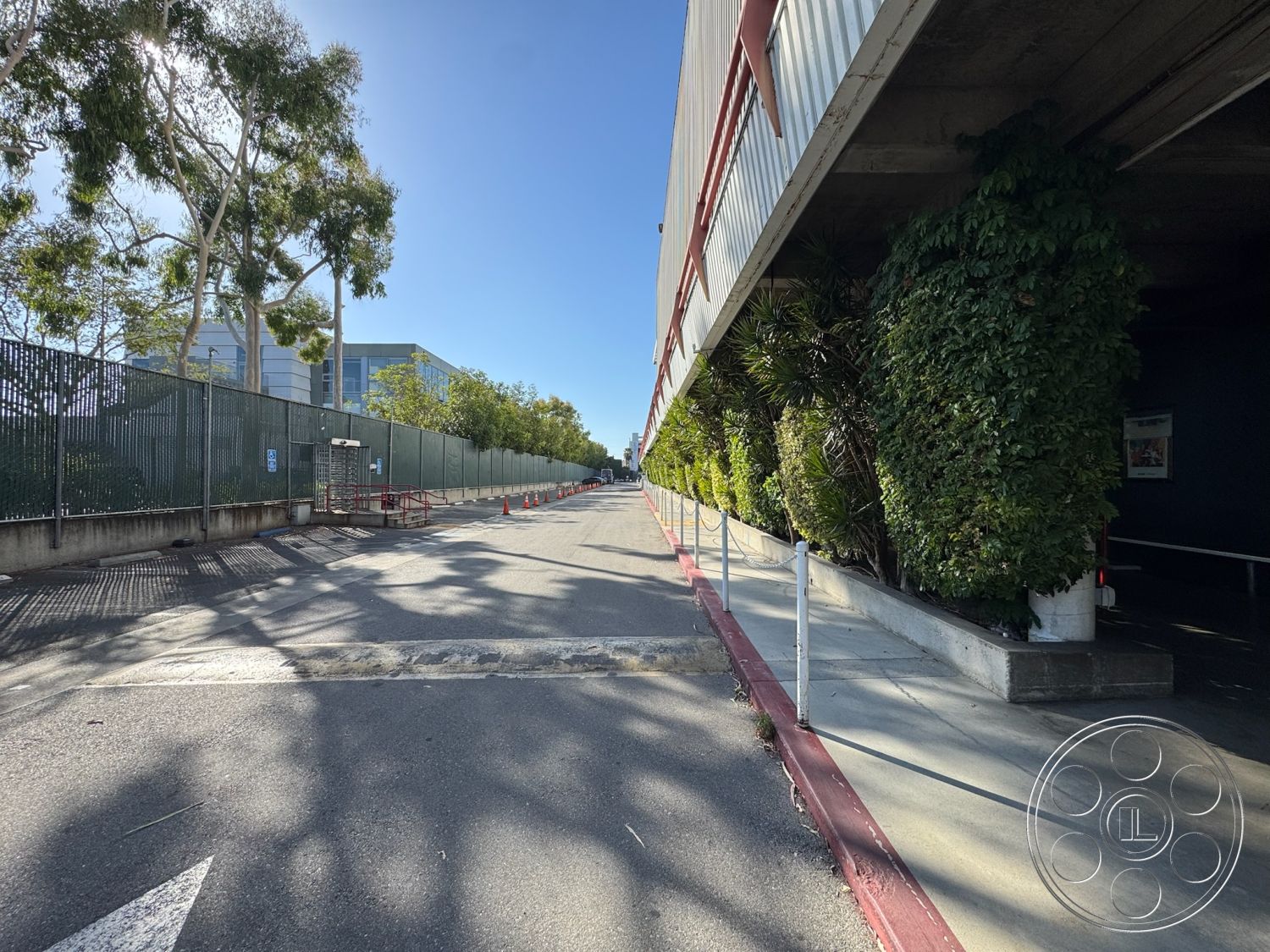 Stage 33 - urban setting, concrete parking surface, exposed concrete structure, curved sidewalk edges, shadow patterns on pavement, landscaped green borders, bright blue sky background, linear planting beds, metal panel exterior cladding, sloped driveway entry, commercial exterior landscaping, tall trees along the boundary, metal fence with chain link, interior-exterior transition areas