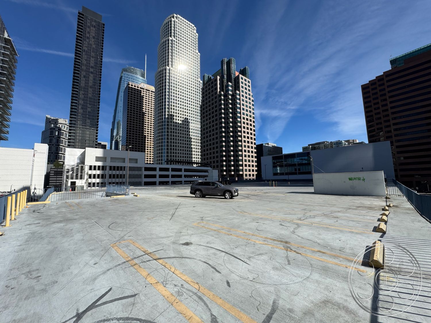 Rooftop 6 - city skyline view, flat roof design, modern architectural elements, urban landscape setting, exposed concrete parking lot surface, concrete parking deck, striped parking markings, metal guardrail details, high-rise urban environment, open-air rooftop parking structure, surrounding skyscraper architecture, exterior urban context, industrial-style structural features, concrete barrier delineation, reflective glass building surfaces