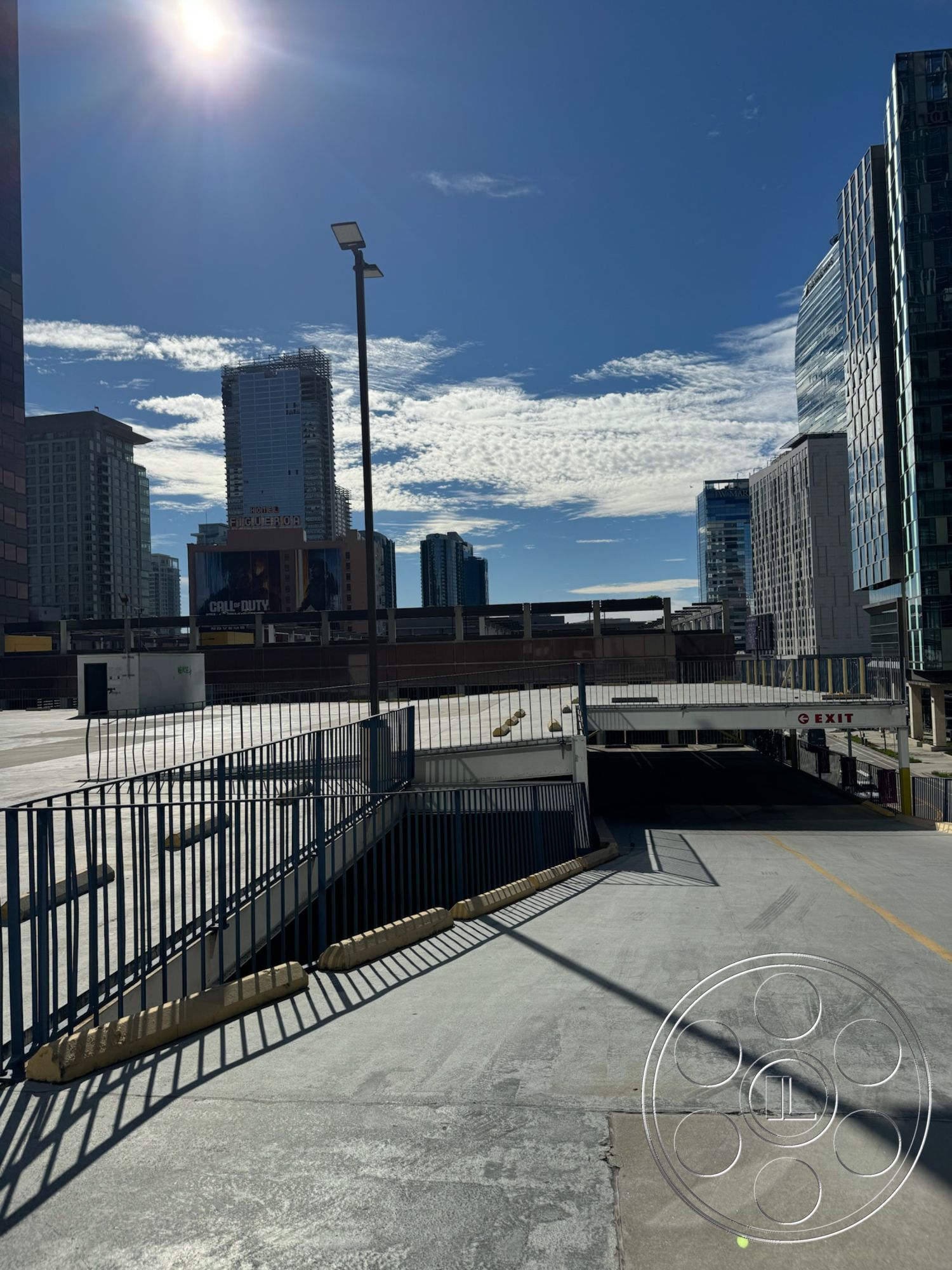 Rooftop 6 - urban setting, city skyline view, clear blue sky, stainless steel fixtures, exterior parking garage structure, concrete parking deck, built-in lighting fixtures, bright sunlight casting shadows, modern high-rise buildings, metal guardrails, sloped driveway leading to underground exit, alternative access ramp, painted concrete surfaces, structured exit signage