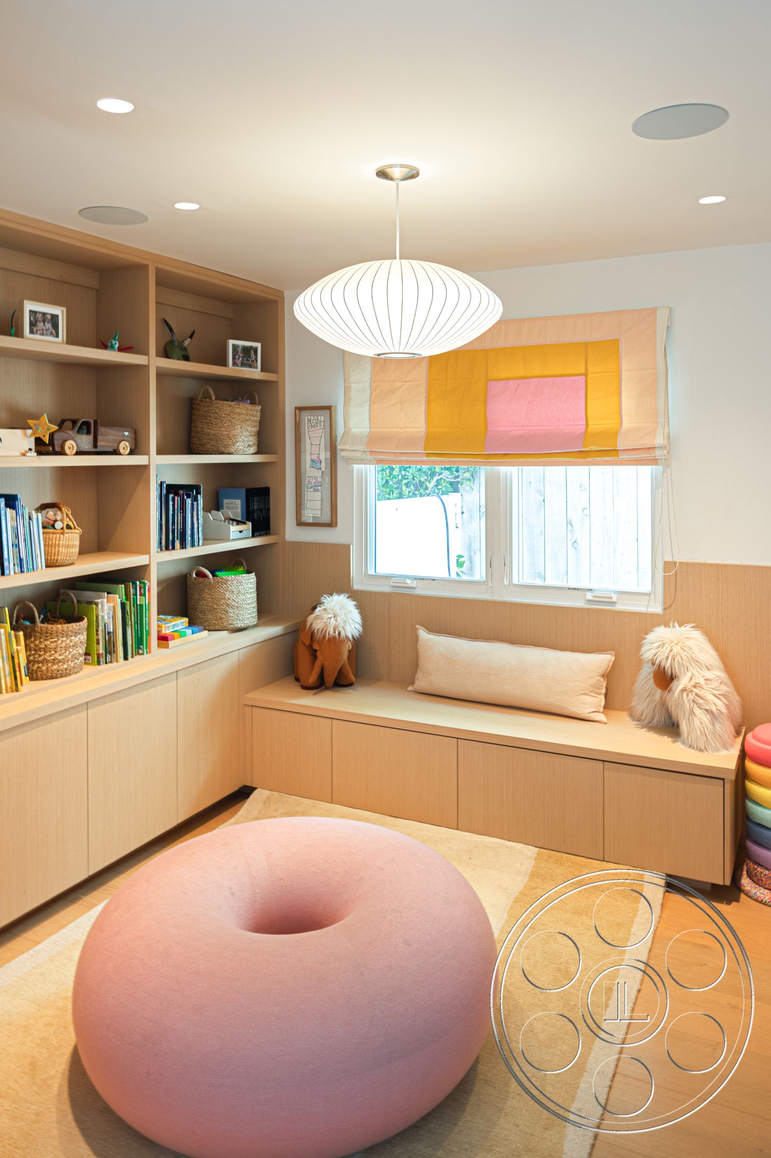 Modern 519 - window view, textured wall treatment, cozy reading nook, child-friendly design, playful accents, minimalist pendant lighting, natural light emphasis, neutral carpet flooring, light wood shelving units, soft pink round ottoman, large window with fabric roller shades, built-in storage bench, decorative woven baskets, colorful children's books