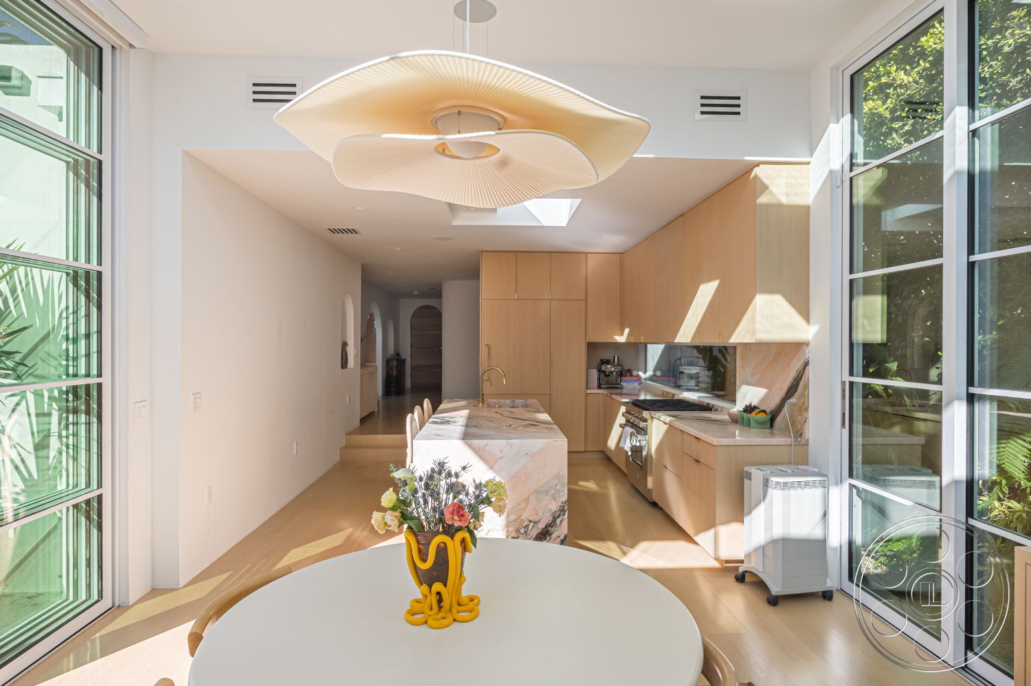 Modern 519 - minimalist design elements, marble kitchen island countertop, interior kitchen with wood cabinetry, lush greenery visible through windows, modern pendant lighting fixture, shiplap accent wall, large glass windows for natural light, light hardwood flooring in dining area, open floor plan with adjacent living space, bright interior with neutral color palette, contemporary dining table with round shape, light-filled space with skylight, smooth surface cabinetry, single-level interior layout