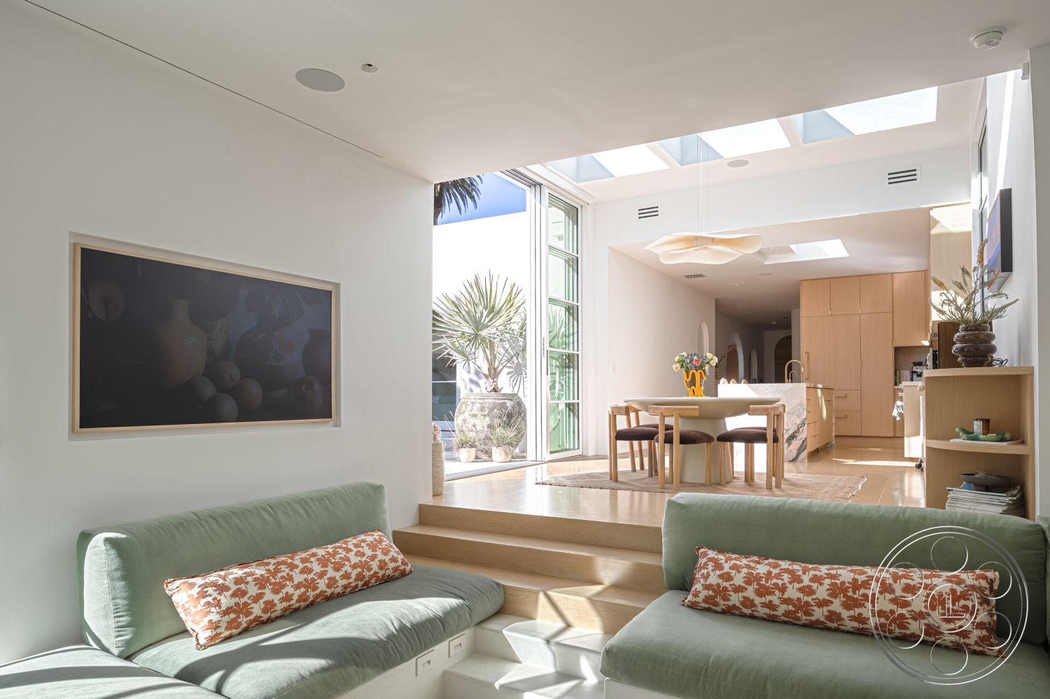 Modern 519 - open floor plan, stainless steel kitchen appliances, marble kitchen island countertop, neutral painted walls, natural light-filled room, area rug under dining table, framed art on wall, interior contemporary living room, green upholstered sofa with patterned throw pillows, wood panel dining table, light wood flooring in living area, skylights allowing natural light, sliding glass doors to outdoor patio, decorative vases with greenery, minimalist cabinetry in kitchen, potted plants by window