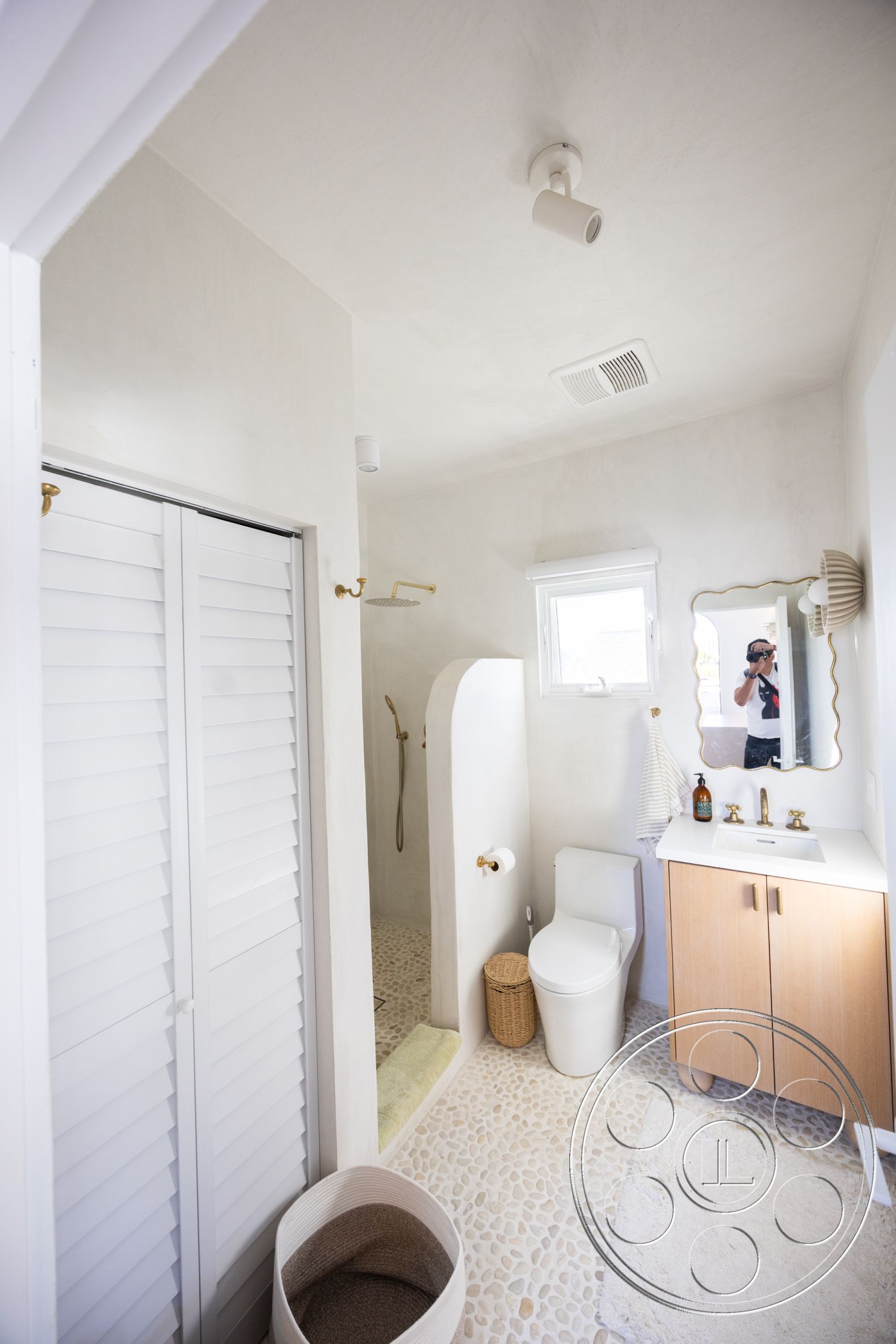 Modern 519 - soft neutral color palette, glass shower enclosure, textured plaster walls, modern minimalist design, interior bathroom, wood vanity cabinet, white painted ceiling, natural light from window, woven storage basket, gold hardware fixtures, pebble shower floor, shuttered storage doors, round mirror above sink, curved shower wall, stylish waste basket, adjustable recessed lighting