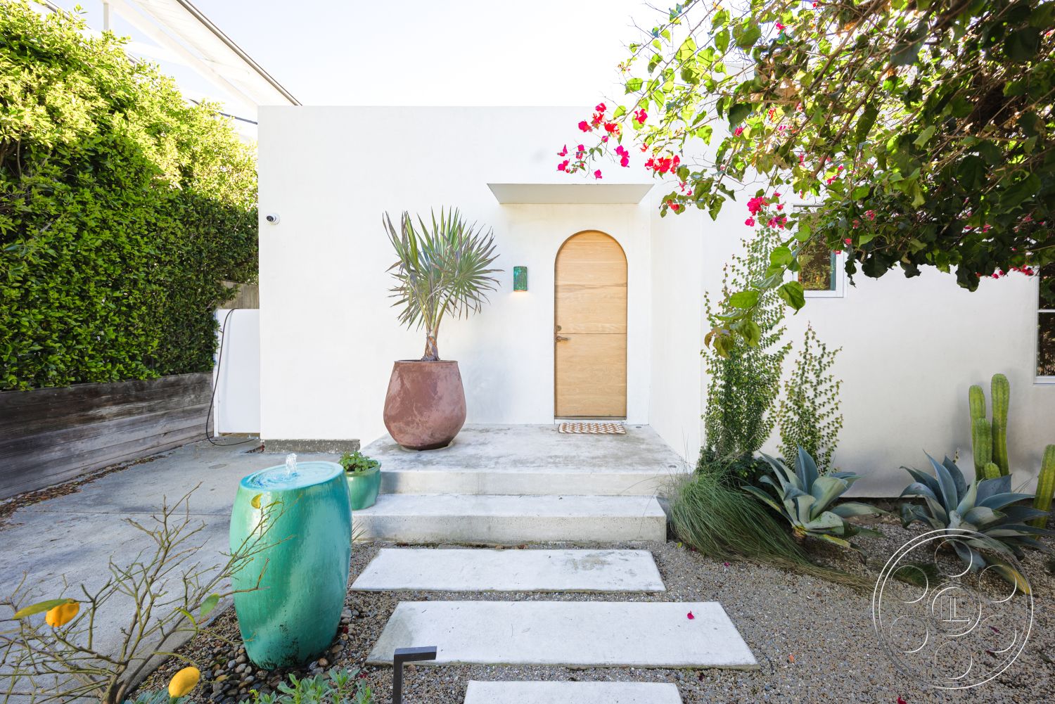 Modern 519 - desert plants, exterior, frontyard, gravel pathway, tropical landscaping, concrete stepping stones, drought-resistant plants, lush greenery backdrop, natural light illumination, outdoor decorative elements, large potted plant, textured outdoor surfaces, modern white stucco walls, arched wooden front door, green ceramic water feature, minimalist house facade, contemporary entrance design