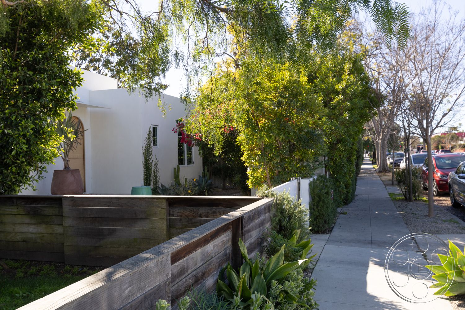 Modern 519 - landscaped garden with native plants, modern residential home, wooden fence exterior, white stucco walls exterior, slate stone paving sidewalk, decorative planter box exterior, hardscaped patio area, arced entryway with wrought iron details, lush foliage surrounding the property, urban residential neighborhood setting, curved pathways through greenery, concrete sidewalk with tree-lined edges, melaleuca trees along the street, designed garden beds, privacy hedges landscaping