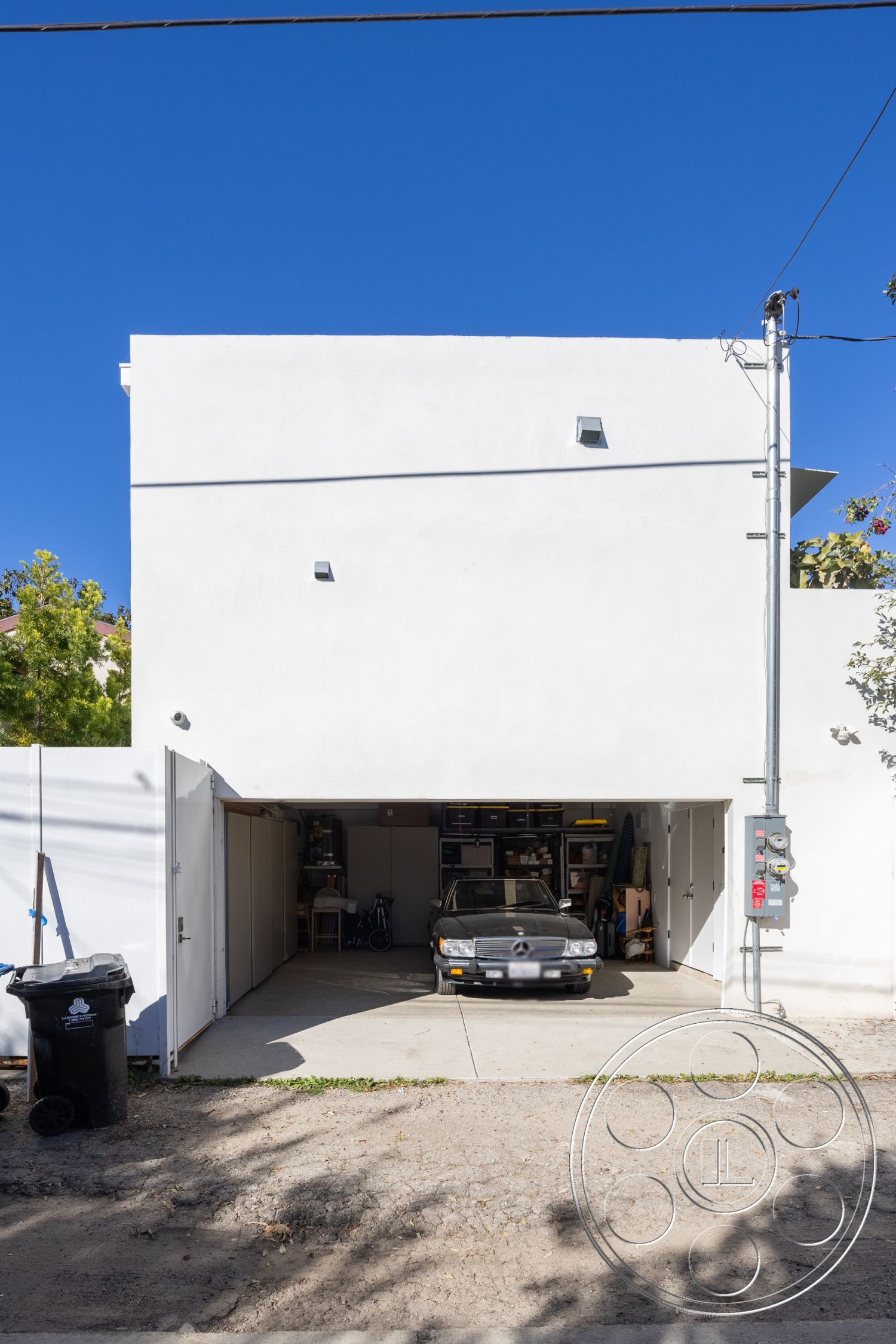Modern 519 - exterior, white stucco walls, concrete driveway, two-car garage, clear blue sky, urban residential area, modern minimalist design, flat roof structure, simple geometric forms, natural stone landscape bordering, garage door with panel design, black asphalt street, plant beds with greenery, utility service box, visual symmetry, visible storage area inside garage
