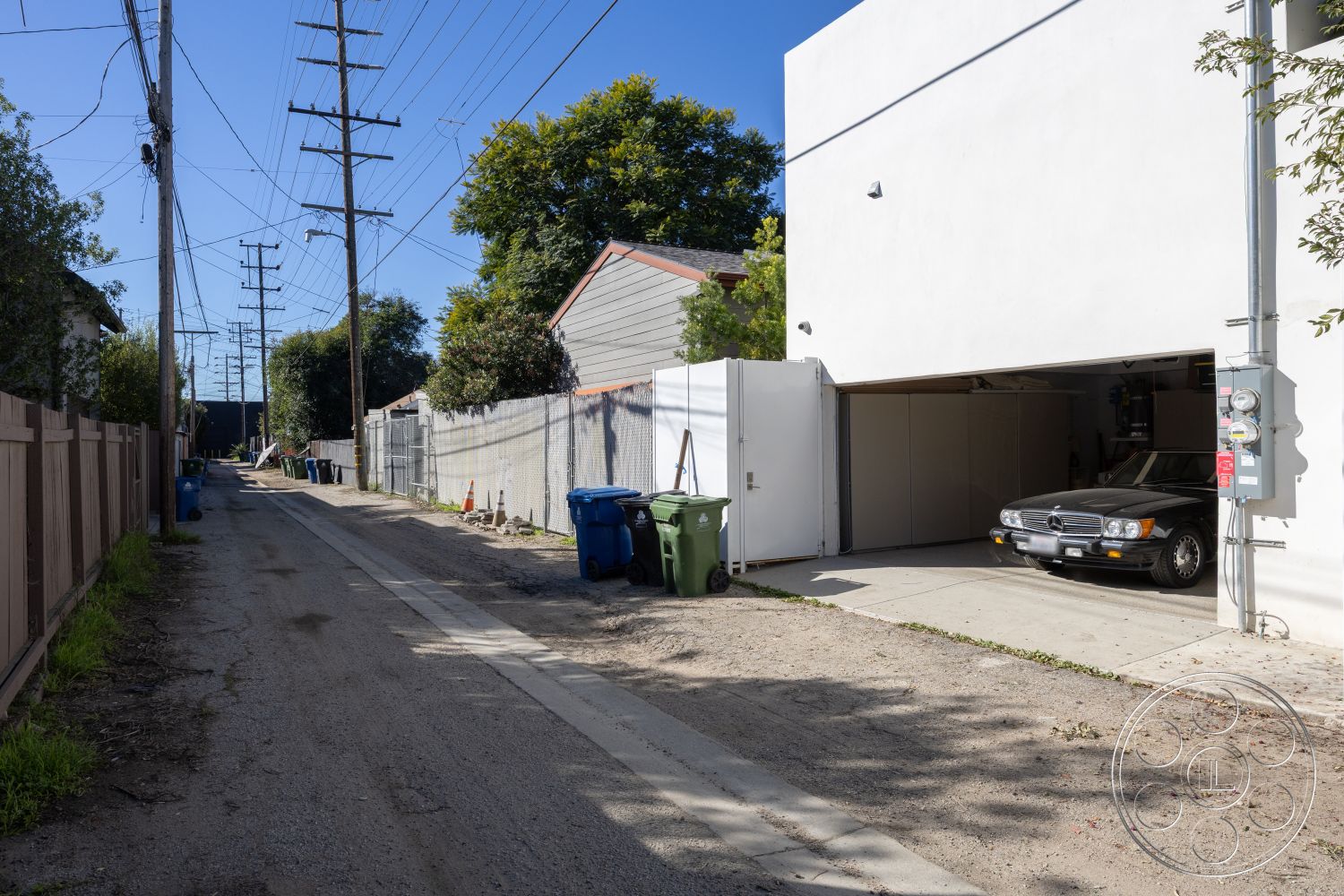 Modern 519 - alley, white stucco wall, residential neighborhood setting, exterior garage door, wooden fence panels, power lines overhead, poured concrete driveway, surrounding gravel alleyway, backyard fence, green recycling bins, black sedan in garage, adjacent residential homes, relaxed urban atmosphere, lush greenery from trees