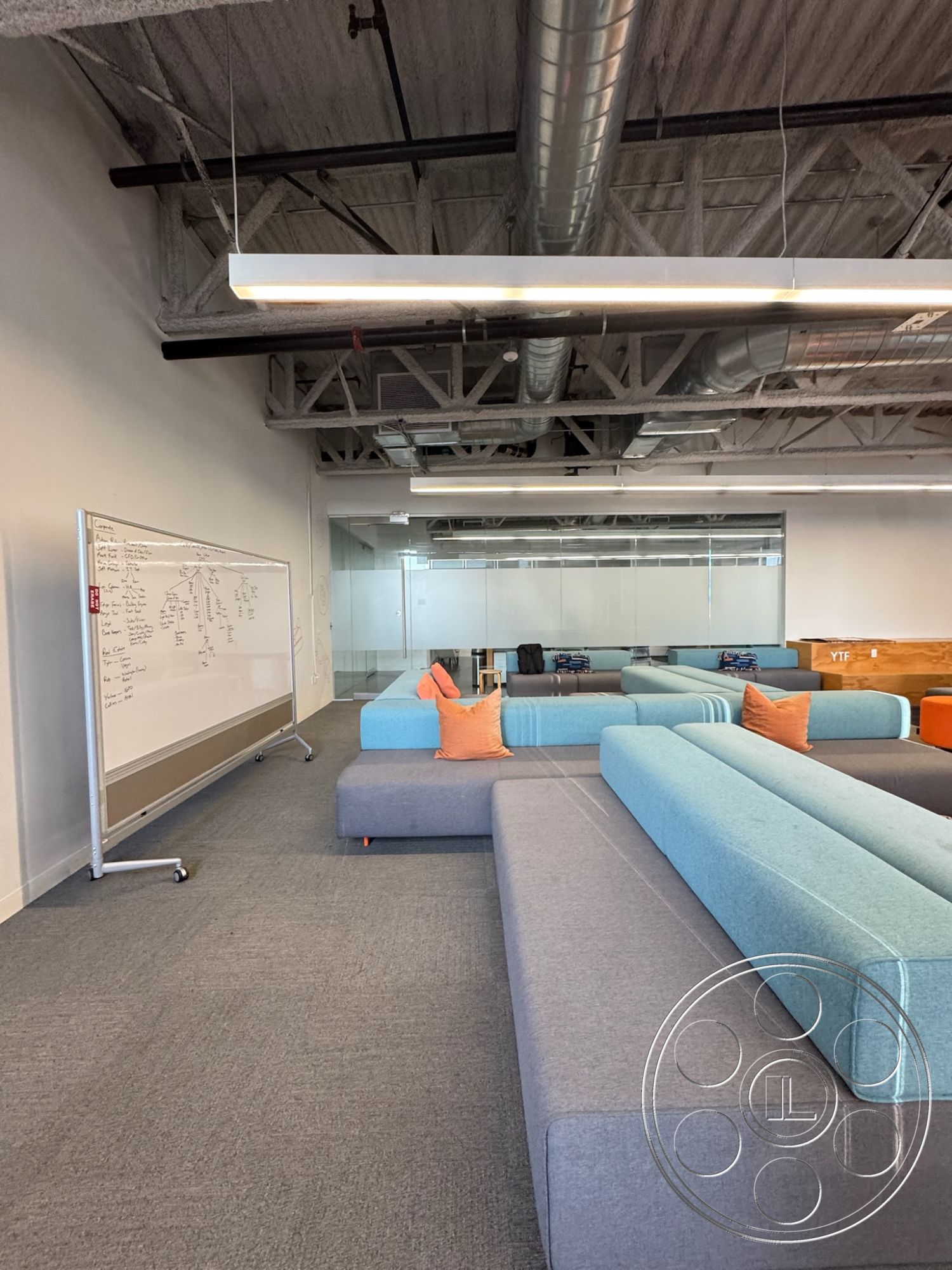 Office 128 - polished concrete flooring, cozy lounge area, glass partition walls, contemporary office design, exposed ductwork ceiling, flexible meeting area, industrial style lighting fixtures, acoustic panel wall treatment, modular seating area, soft grey upholstery couches, bright orange throw pillows, whiteboard for collaboration, open concept office space, urban workplace setting, modern workspace interior