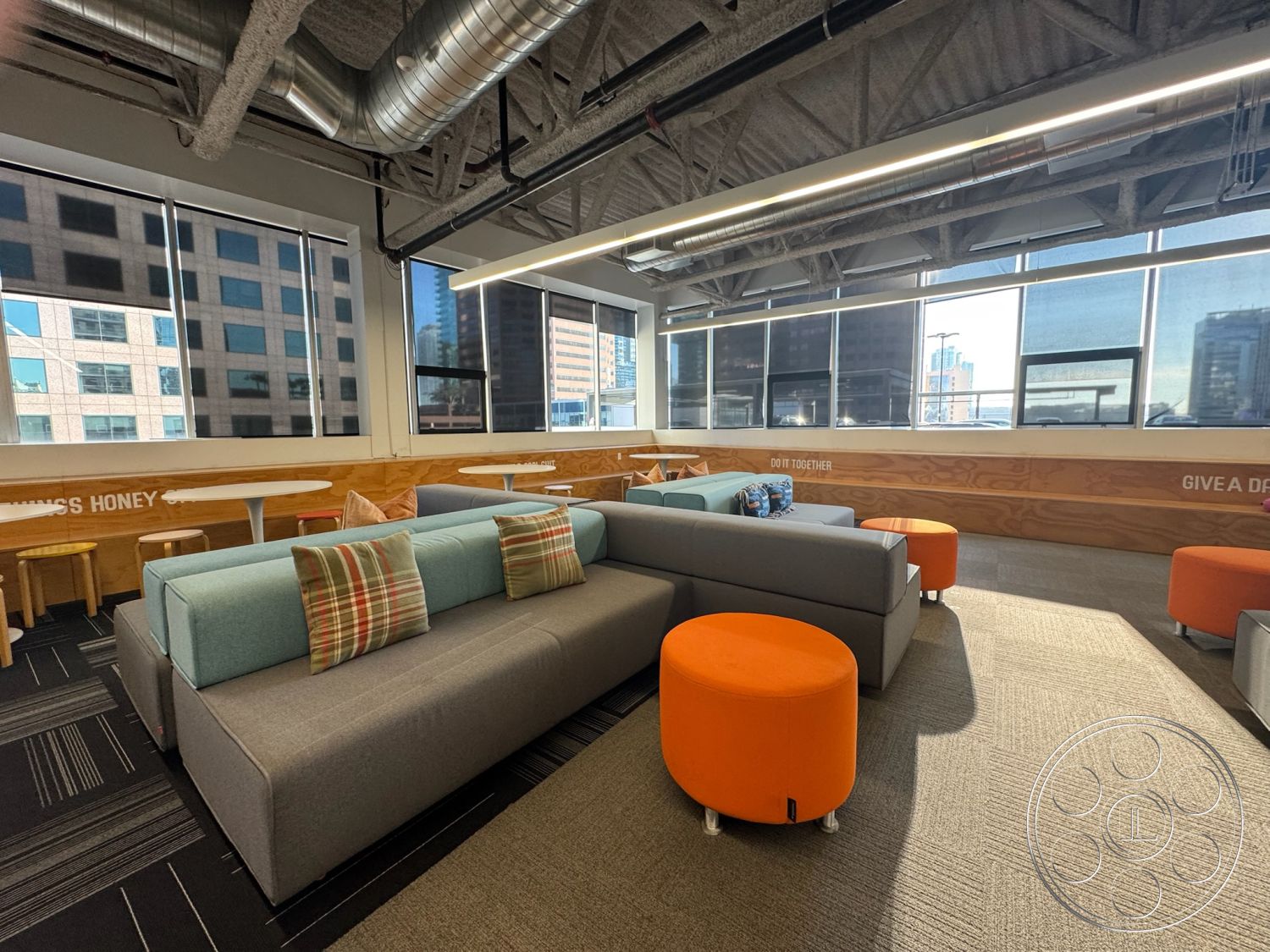 Office 128 - collaborative workspace, large floor-to-ceiling windows, interior lounge area, contemporary office design, exposed ductwork ceiling, light gray carpet flooring, urban city skyline view, soft seating arrangements, modular gray upholstery sofa, plaid decorative pillows, round orange side tables, wood accent wall paneling, bright and inviting atmosphere