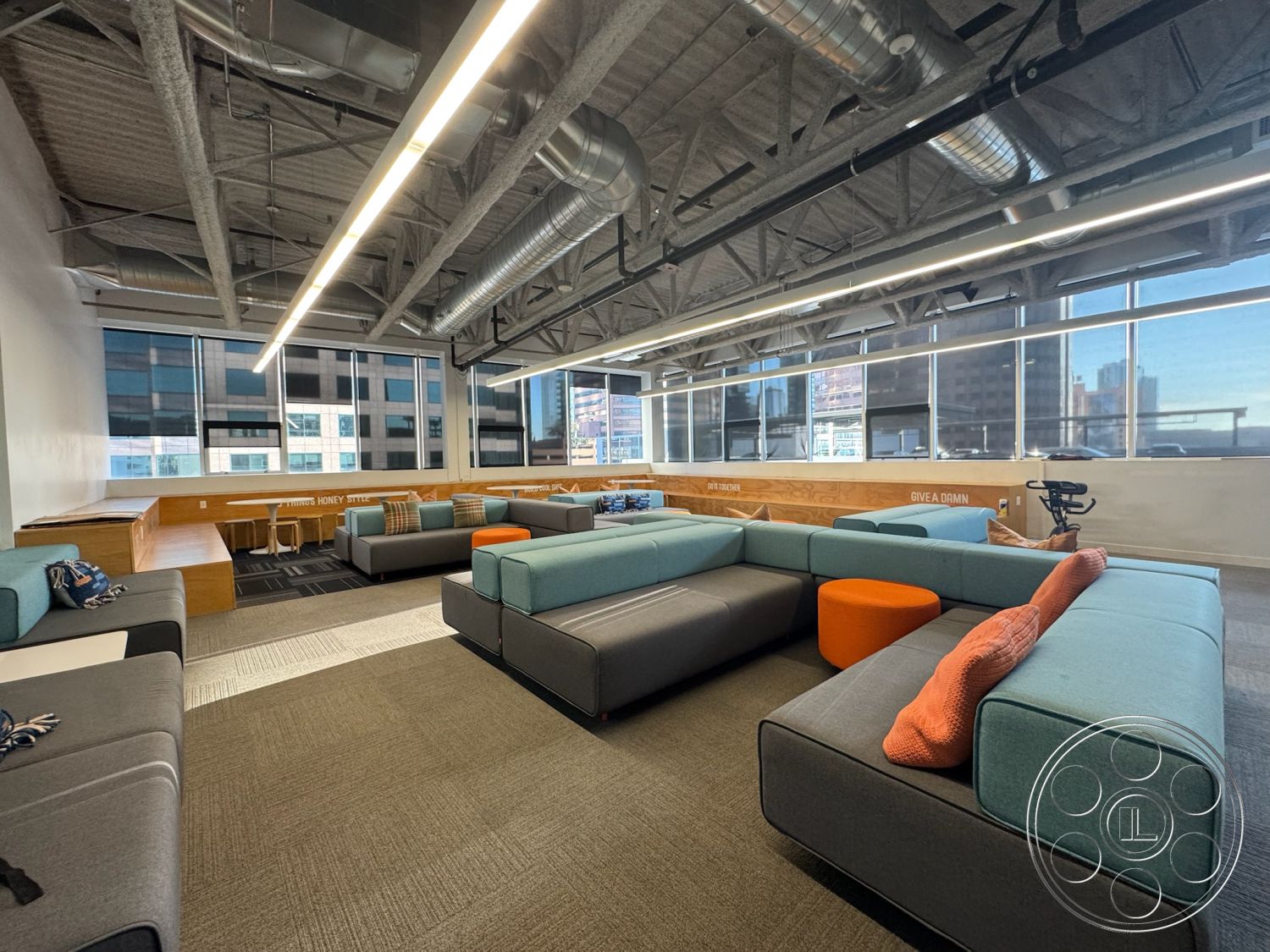 Office 128 - textured wall panels, wooden bench seating, casual meeting area, exposed concrete ceiling, gray carpet flooring, view of city skyline, glass partition walls, interior lounge space, LED ceiling lighting, open layout office, large panoramic windows, industrial-style design, modular fabric seating, multicolored accent cushions, collaborative workspace lounge