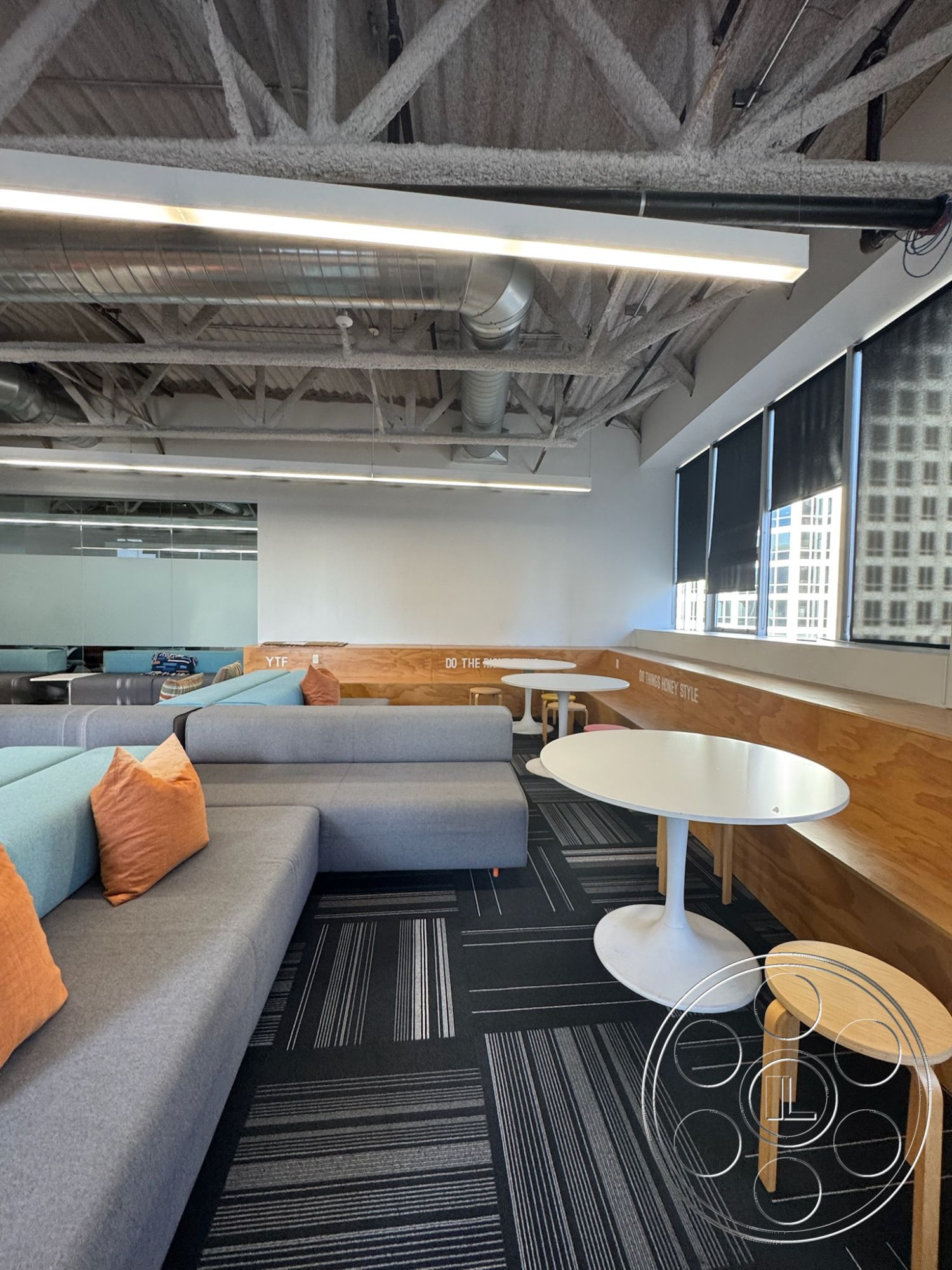 Office 128 - collaborative workspace, floor-to-ceiling windows, contemporary design elements, open office layout, textured carpet flooring, industrial-style interior, exposed metal ceiling beams, wood panel wall treatment, grey upholstered sectional sofa, soft seating lounge area, blue accent cushions, white round dining table, light wood circular table base, striped pattern carpet