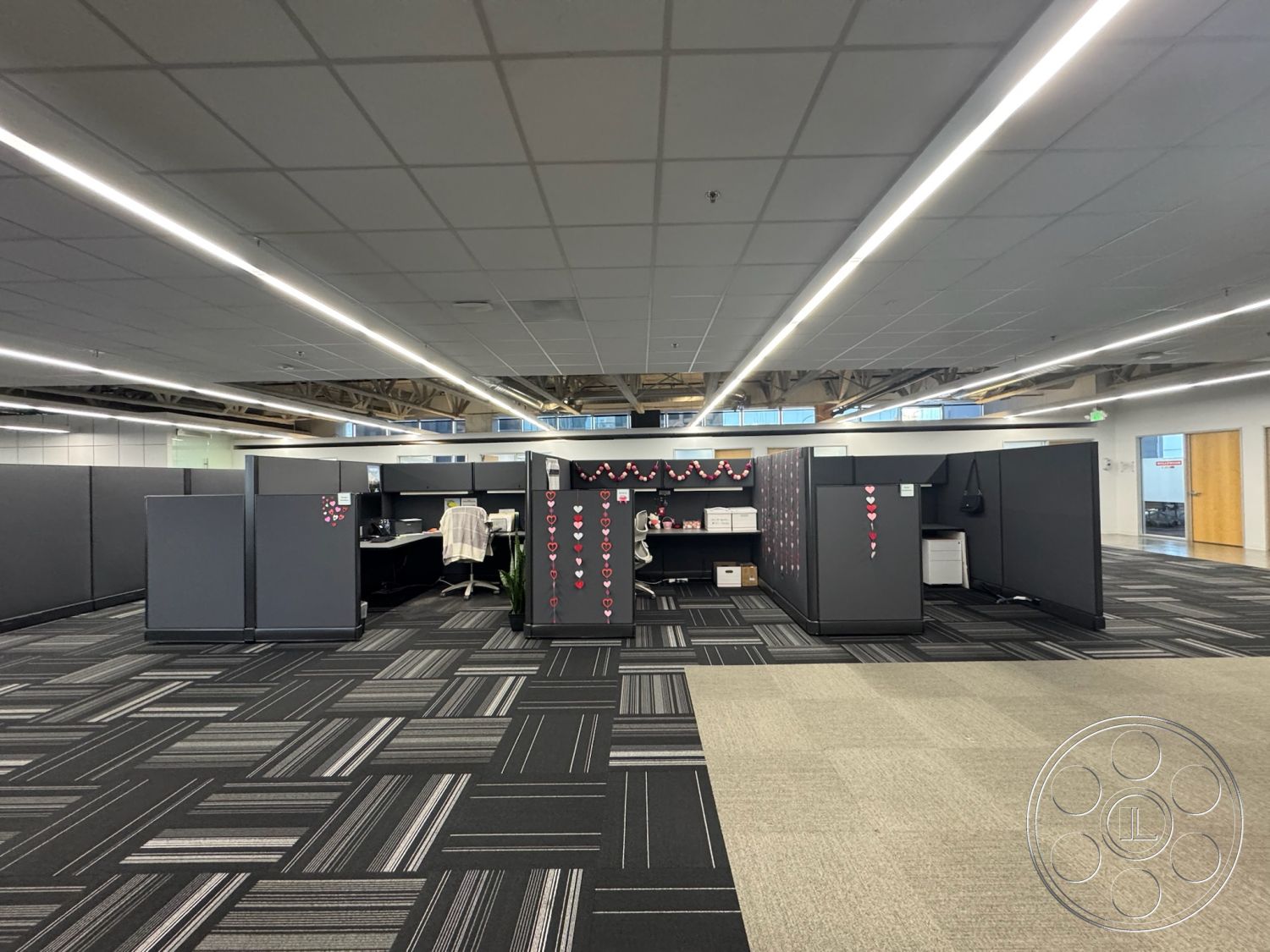 Office 128 - neutral color palette, open-plan layout, faux plant decor, large windows with natural light, interior office space, fluorescent ceiling lights, decorative wall hangings, organized shelving units, high ceiling with exposed beams, modular cubicles, gray fabric partition walls, black and white carpet tiles, workstation with ergonomic chair, entry door with wood finish, collaborative workspace area