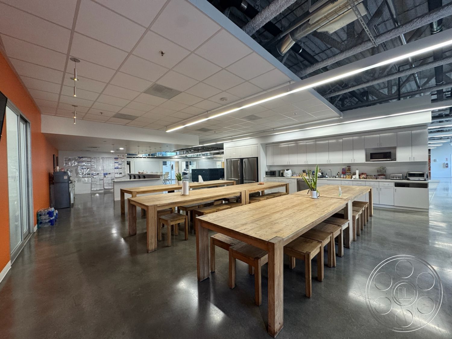 Office 128 - polished concrete flooring, functional workspace, natural light from large windows, modern kitchen island, pendant lighting fixtures, interior dining area, exposed ductwork ceiling, industrial-style lighting, commercial kitchen area, open-plan kitchen design, large wooden dining tables, simple wood dining chairs, white cabinetry with stainless steel appliances, minimalist space, bright orange accent wall, contemporary office break room
