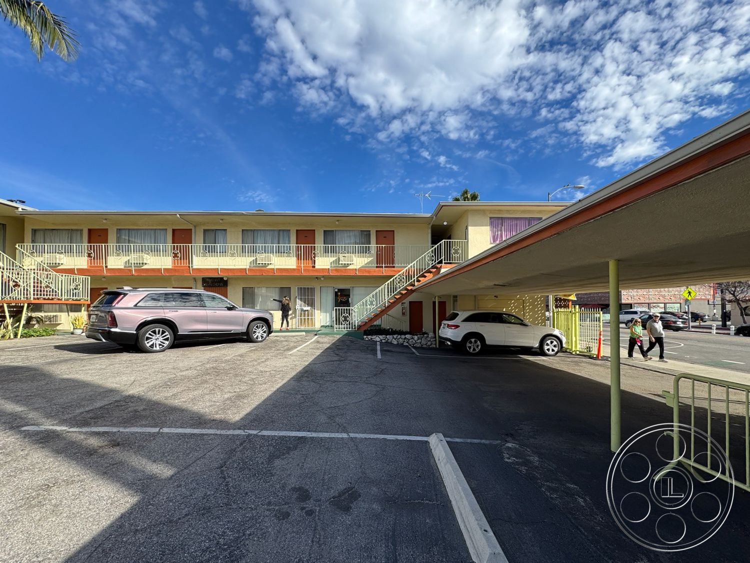 Hotel 59 - exterior, cream stucco walls, flat roof design, palm tree landscaping, metal stair railings, balcony overhang, sunny blue sky, urban motel setting, composite parking area, paved asphalt parking lot, colored door accents, second-story walkway, cars in parking, open parking structure, ground-level carports