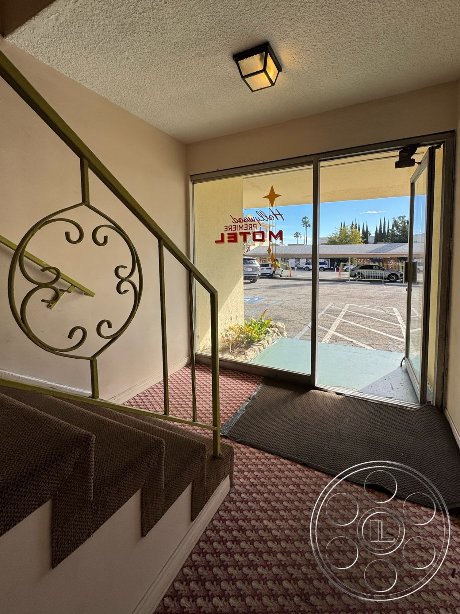 Hotel 59 - bright natural lighting, carpeted stairs, pendant lighting fixture, neutral painted walls, metal stair railings, glass door entryway, textured ceiling finish, welcoming reception area, potted plants at entrance, patterned carpet flooring, interior entrance area, vintage motel decor, exterior parking lot view, open doorway to exterior, arched entryway design