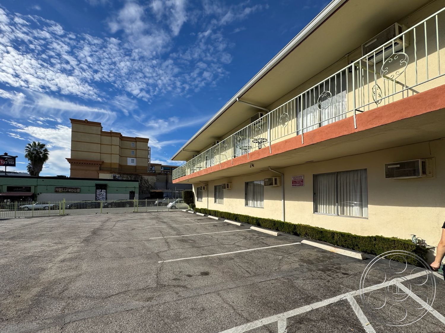Hotel 59 - exterior, urban setting, surrounding buildings, commercial area, balcony with wrought iron railings, concrete parking lot, city backdrop, sunny clear sky, palm tree in foreground, two-story motel structure, red accent trim, stucco hotel facade, air conditioning units on wall, striped asphalt parking lines, landscaped shrubbery along perimeter, large windows with sheer white curtains