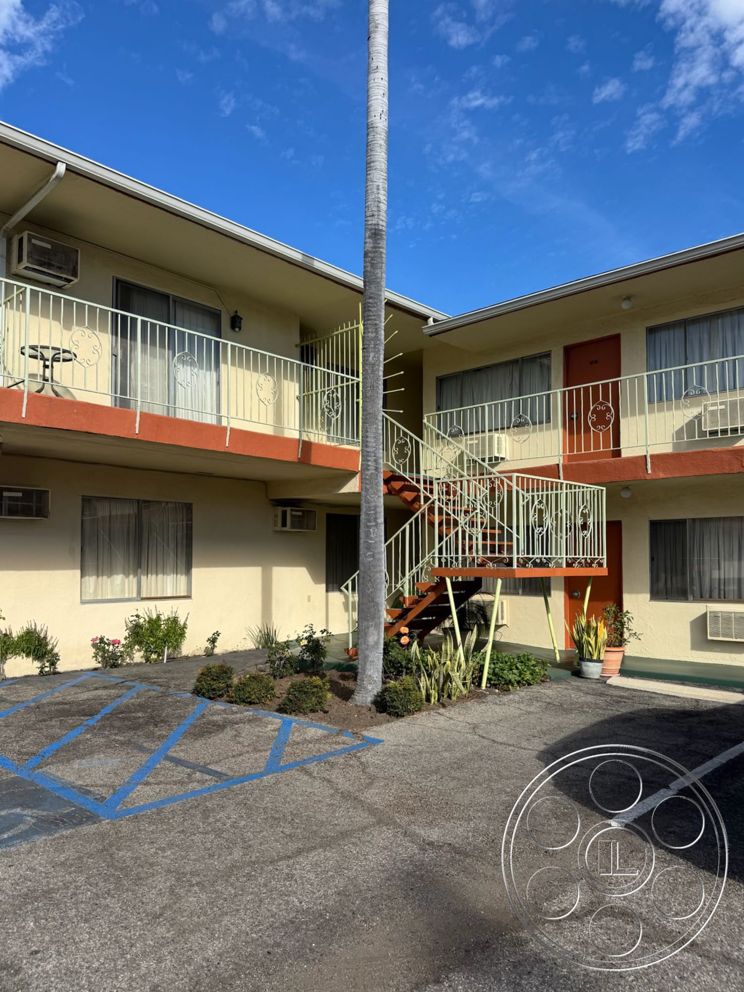 Hotel 59 - urban setting, clear blue sky, functional outdoor space, exterior landscaped garden beds, exterior concrete parking lot, exterior concrete steps, exterior palm tree landscaping, exterior painted stucco walls, exterior asphalt driveway, exterior metal balcony railing, exterior wooden entrance door, exterior spiral staircase, exterior glass sliding windows, exterior blue handicap parking markings, two-story apartment building, courtyard-style layout