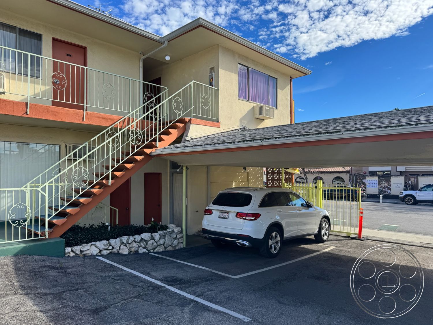 Hotel 59 - exterior, urban setting, gravel landscaping, terracotta roof tiles, concrete parking lot, curved balcony railing, outdoor patio area, metal stair railing, rectangular concrete steps, modern carport design, two-story apartment building, yellow stucco exterior walls, red painted doors, built-in parking area, white stone garden border, cloudy blue sky