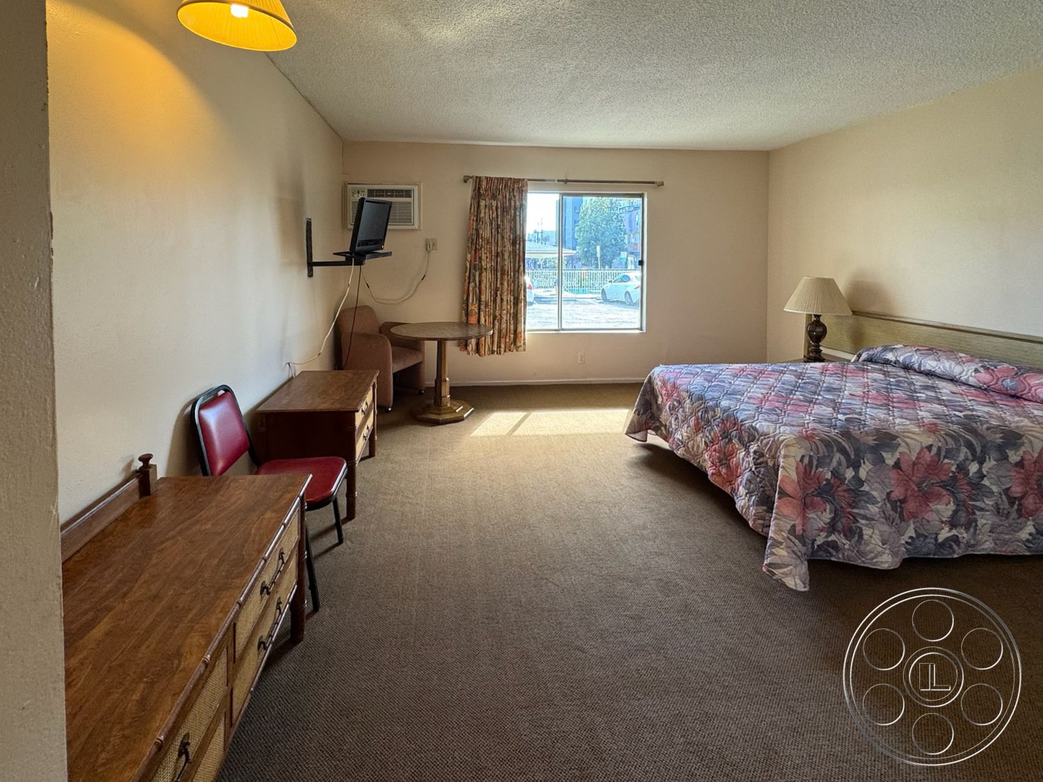 Hotel 59 - wall-mounted television, textured ceiling, window with natural light, wooden side table, round wooden coffee table, carpet bedroom flooring, light beige painted walls, red upholstered chair, floral patterned bedding, wooden dresser with multiple drawers, curtains with floral pattern, chair with metal frame