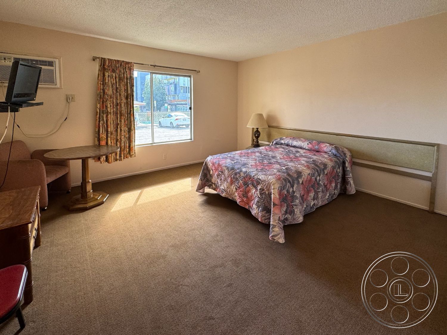 Hotel 59 - natural light, wooden coffee table, floor-to-ceiling window, upholstered armchair, simple bedside lamp, brown wooden furniture, neutral painted walls, minimalist interior, carpet bedroom flooring, lampshade table lamp, plush floral bedspread, curtain window treatment, wall-mounted air conditioning unit, guest bedroom