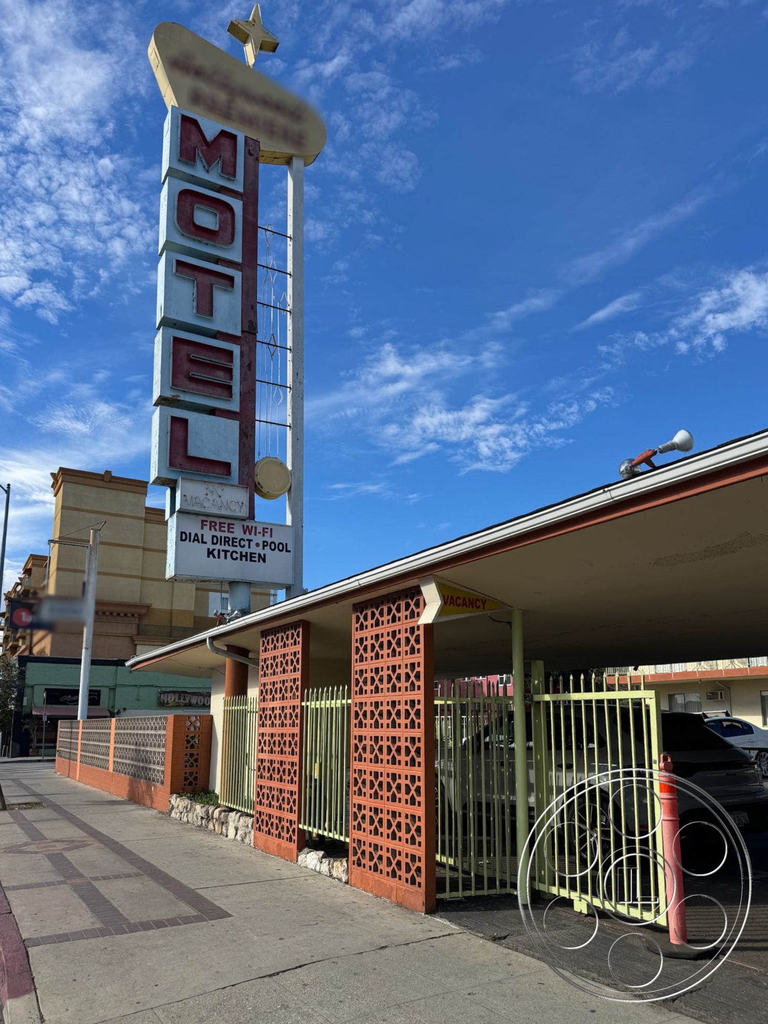Hotel 59 - exterior, paved driveway, mid-century modern architectural style, sunny day setting, commercial building facade, exterior lighting fixtures, urban motel environment, retro motel sign, blue metal sign with white lettering, brick wall with decorative cutouts, orange concrete accents, green metal fencing, stone planter wall, vacant parking space, covered parking area, amenities sign with travel information
