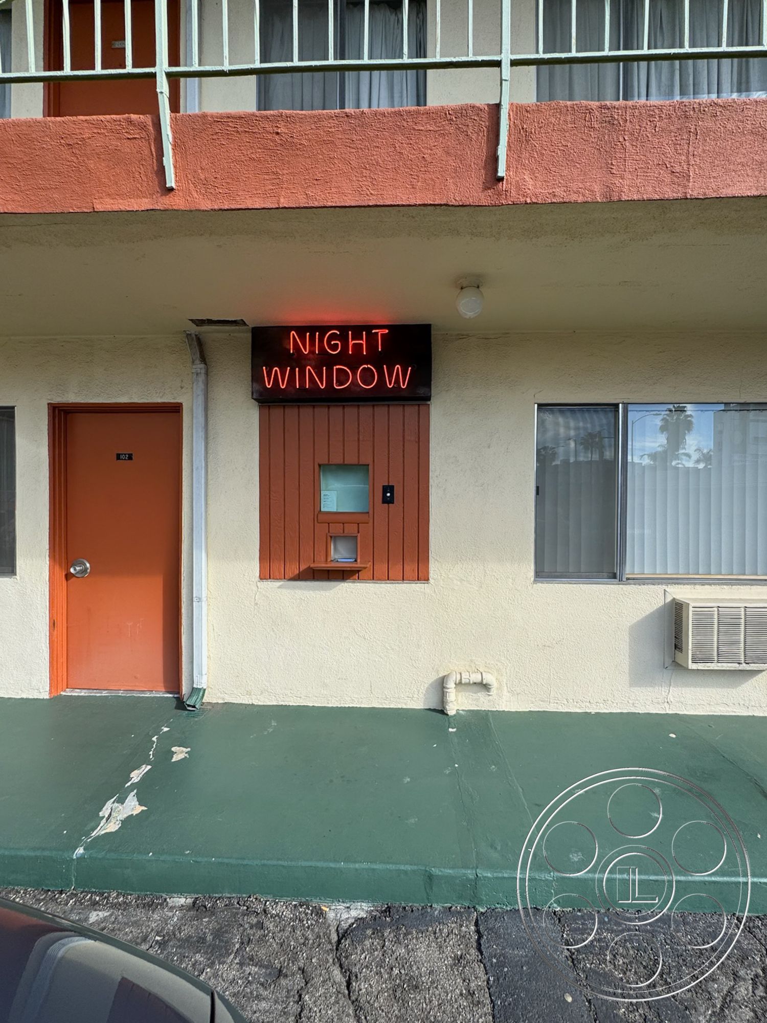 Hotel 59 - exterior, minimalist design elements, urban setting, air conditioning unit, stucco wall finish, metal railing balcony, bright orange door, green concrete floor, wood panel accent around window, neon sign, single-storey motel layout, horizontal window treatments, night window service, motel aesthetic