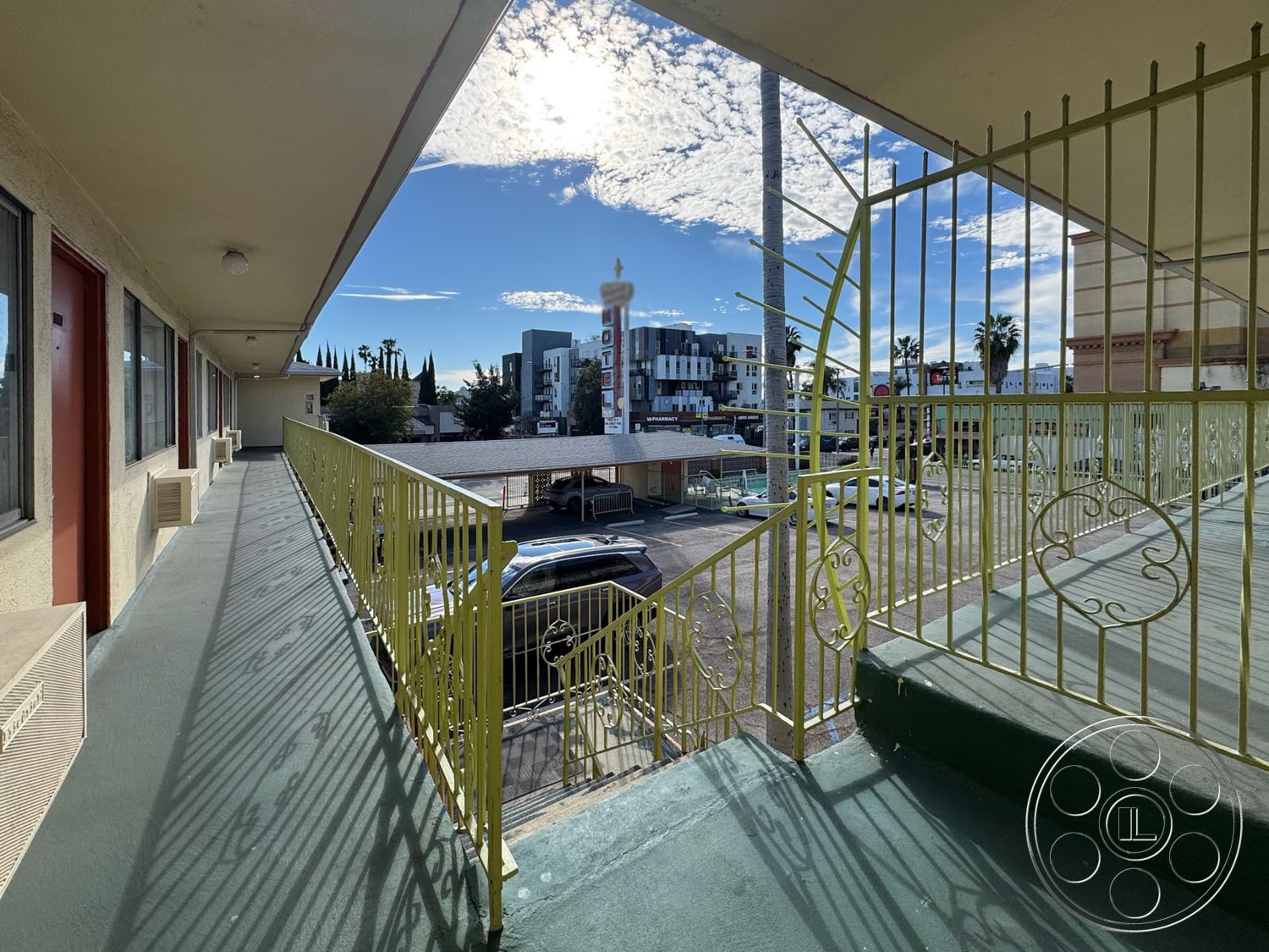 Hotel 59 - palm tree view, stucco wall finish, flat roof structure, white painted window frames, urban hotel setting, open-air balcony design, concrete walkway floor, exterior corridor with metal railing, yellow powder-coated metal stair railings, wooden door with red paint, outdoor motel balcony, modern city skyline view, multi-story building architecture, indoor air conditioning unit