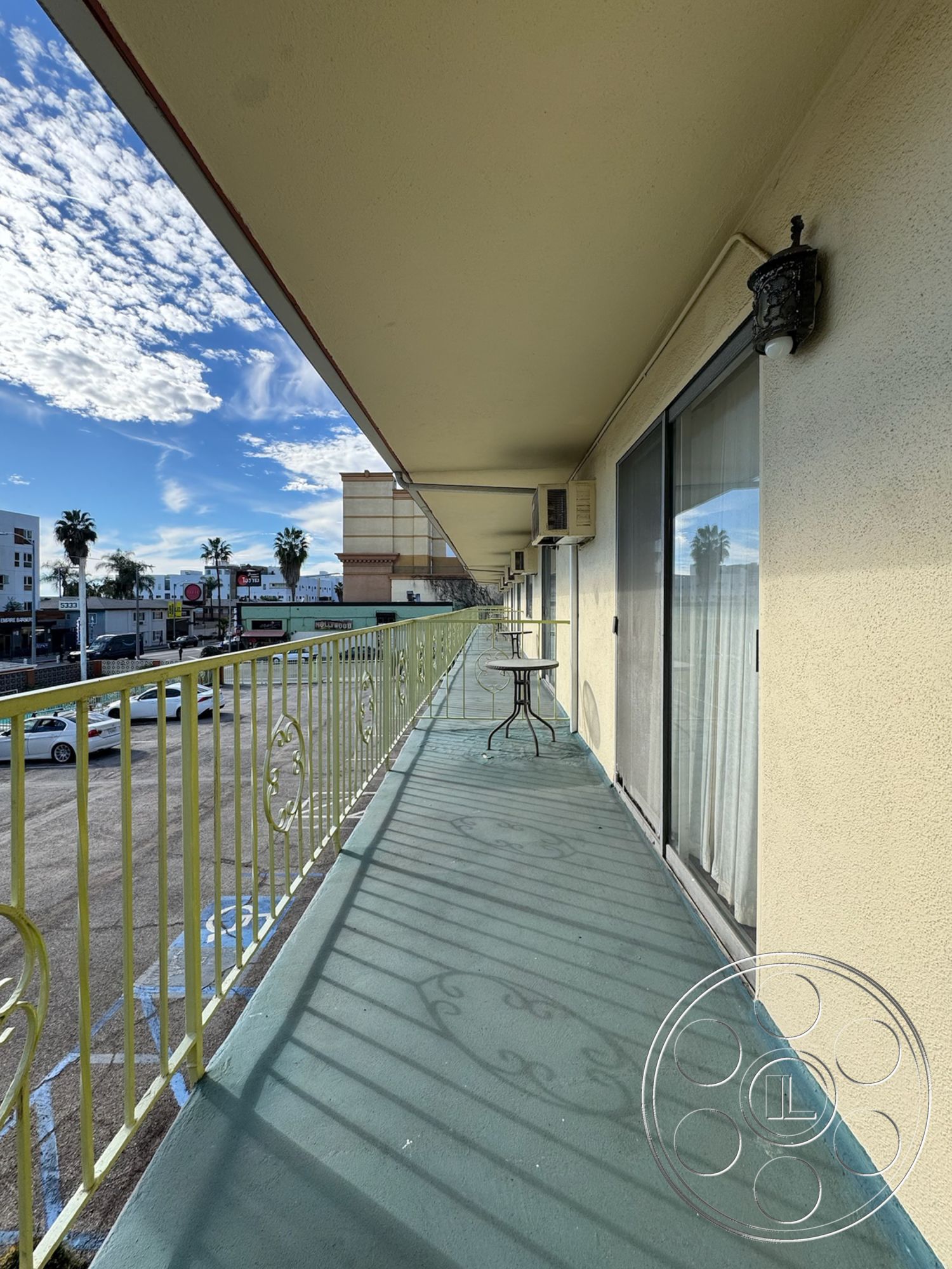 Hotel 59 - natural lighting, cityscape view, urban environment, exterior balcony, palm tree landscape, two-story building structure, shadow patterns on floor, concrete balcony flooring, yellow wrought iron railings, rectangular exterior space, textured beige wall surfaces, small round table and chair setup, attached to hotel-style building