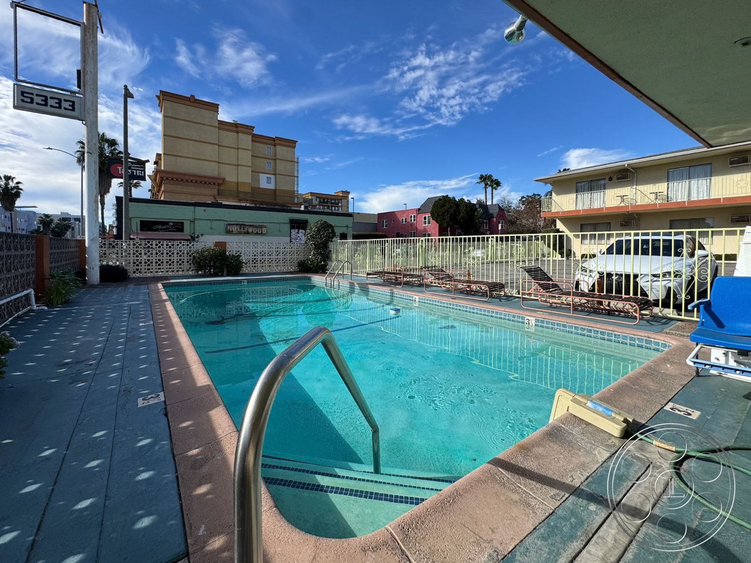 Hotel 59 - steel railing, city skyline view, clear blue sky, poolside loungers, rectangular pool, wooden lounge chairs, sunny outdoor space, surrounding palm trees, exterior concrete pool border, concrete patio flooring, concrete parking area, textured concrete deck, metal pool fencing, suburban motel setting, painted brick wall, decorative block wall