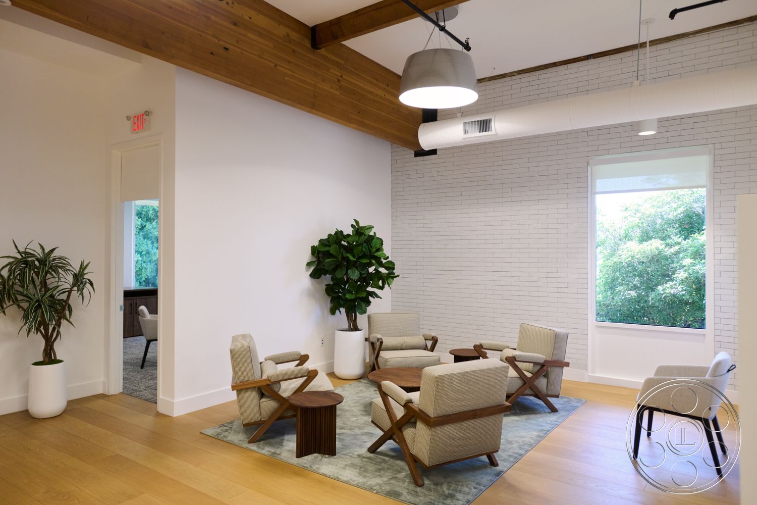 Office 129 - textured area rug, light hardwood flooring, round wooden coffee table, modern lounge area, exposed wood ceiling beams, interior seating area, natural light interior, white painted walls, minimalist furniture design, indoor potted plant, contemporary workspace, large picture windows, upholstered accent chairs, industrial ceiling light fixture