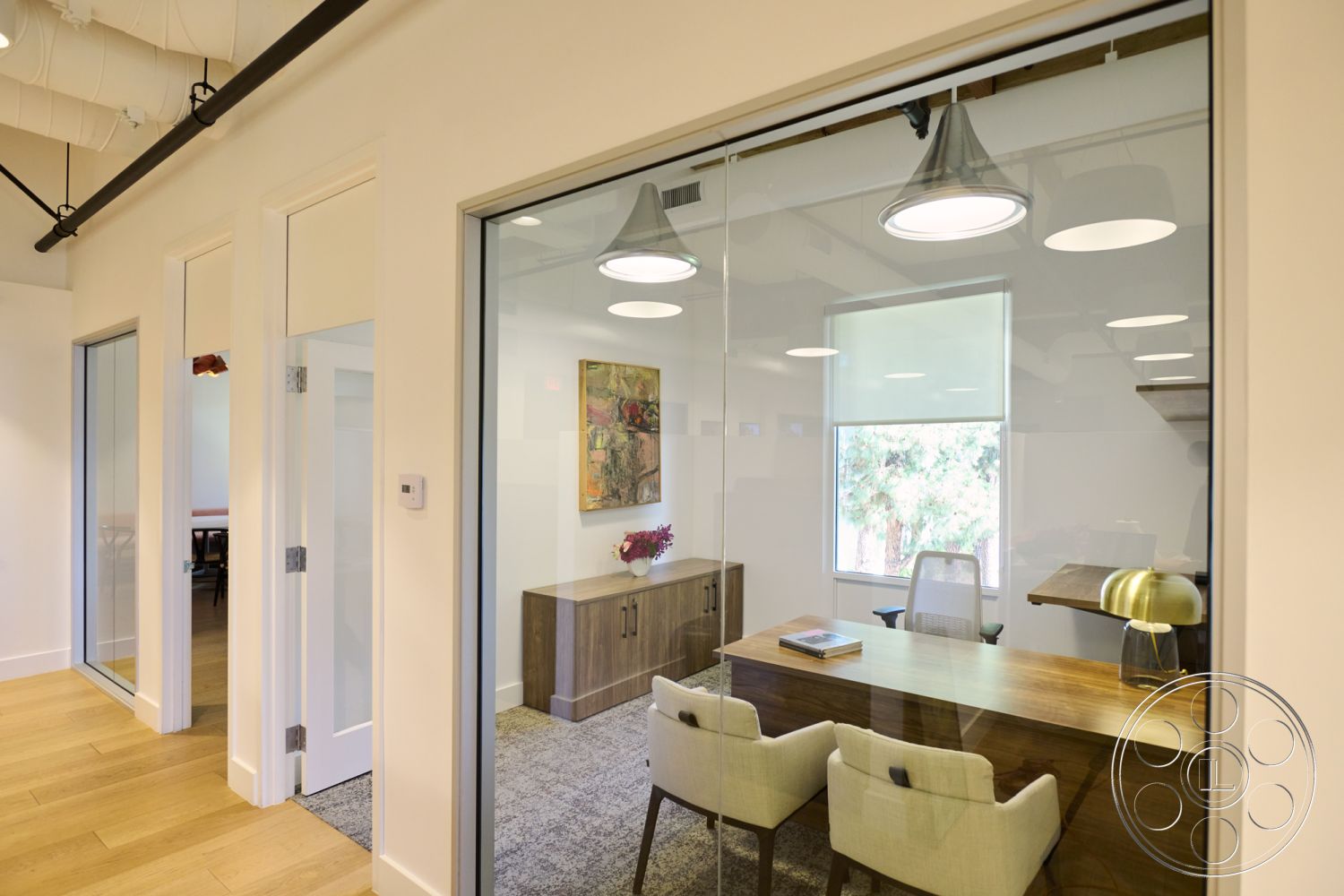 Office 129 - neutral color palette, hardwood flooring, modern light fixtures, minimalist furniture, artwork on the wall, window with natural light, glass partition walls, textured carpet flooring, interior office space, contemporary office design, wooden office desk, open workspace layout, exposed ceiling ductwork, lounge area with two chairs, desk lamp with metallic finish, accent decorative elements