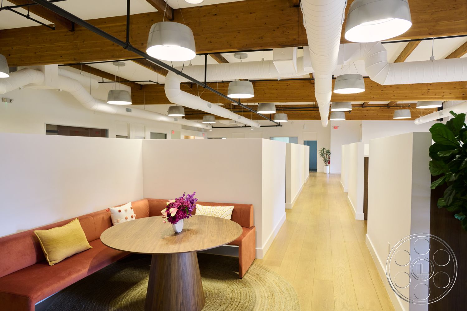 Office 129 - contemporary design elements, natural light from windows, round wooden dining table, exposed wood ceiling beams, neutral color palette with accents, interior modern office space, stylish pendant lighting fixtures, white drywall partitions, vinyl plank flooring, cushioned corner bench seating, bright floral centerpiece, open floor plan office layout, soft carpet area rug, industrial style air ducts, workspace with collaborative seating