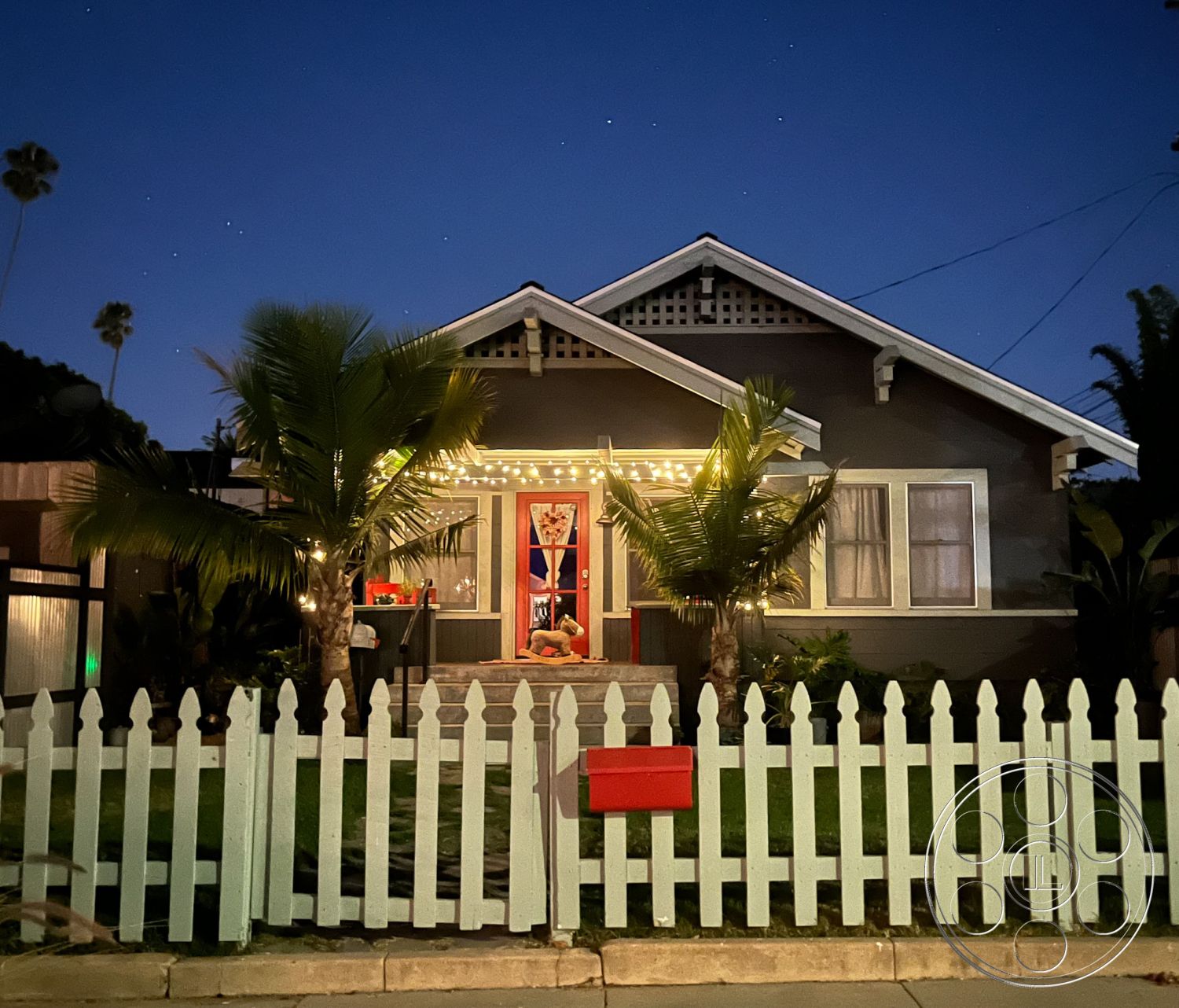 Craftsman 56 - exterior, suburban neighborhood, stone pathway, nighttime setting, gabled roof, urban garden, white picket fence, gray wood siding, green grass lawn, palm tree landscaping, red front door, Craftsman-style home, concrete front steps, decorative eave brackets, outdoor string lights, porch with decorative railing, warm inviting entryway