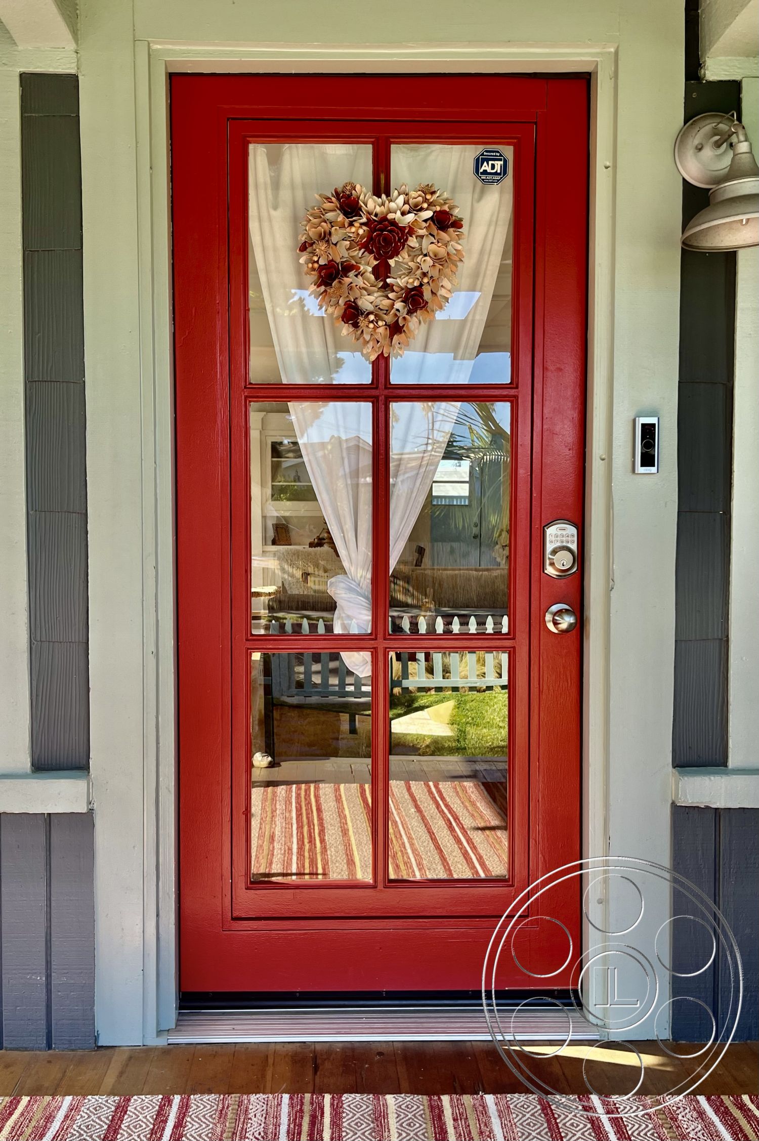 Craftsman 56 - striped outdoor rug, exterior light fixture, metal door hardware, green lawn landscaping, red front door with glass panels, decorative heart-shaped wreath, exterior trim detailing in light gray, wood door frame with vertical panels, gray clapboard siding, wooden porch flooring, traditional porch with railing, interior glimpse of living space, suburban front entryway, curtain drapes on door, picket fence view