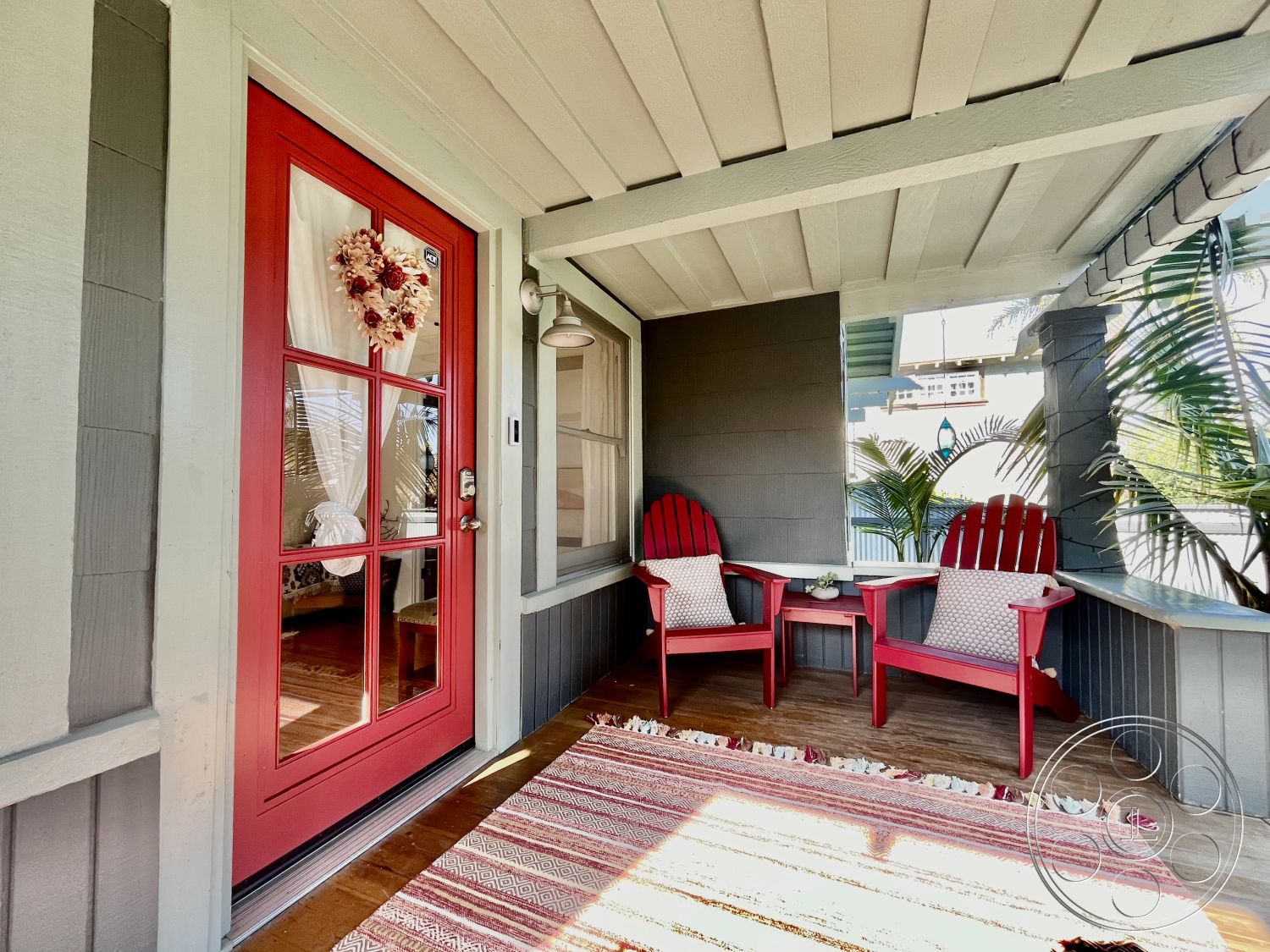 Craftsman 56 - exterior, welcoming entryway, decorative throw pillows, tropical setting, white window trim, woven area rug with geometric pattern, cozy outdoor seating area, gray clapboard siding, wooden porch flooring, red door with window panels, light fixture above seating area, red adirondack chairs, green tropical plants, hanging lantern light fixture, white curtains framing door, porch ceiling with exposed beams, small wood side table, aesthetic porch decor