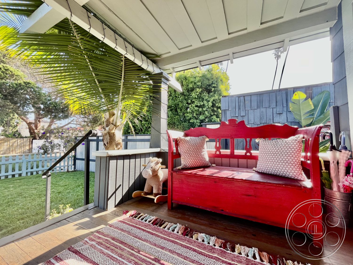 Craftsman 56 - woven area rug, exposed wooden ceiling beams, sunlit outdoor space, urban backyard setting, outdoor potted plants, exterior porch seating area, red painted wood bench with cushions, shiplap wall paneling, green palm tree landscaping, fenced yard with green grass, decorative pillows with geometric patterns, wooden floor decking, cozy relaxation nook, comfortable outdoor living, stylish patio decor