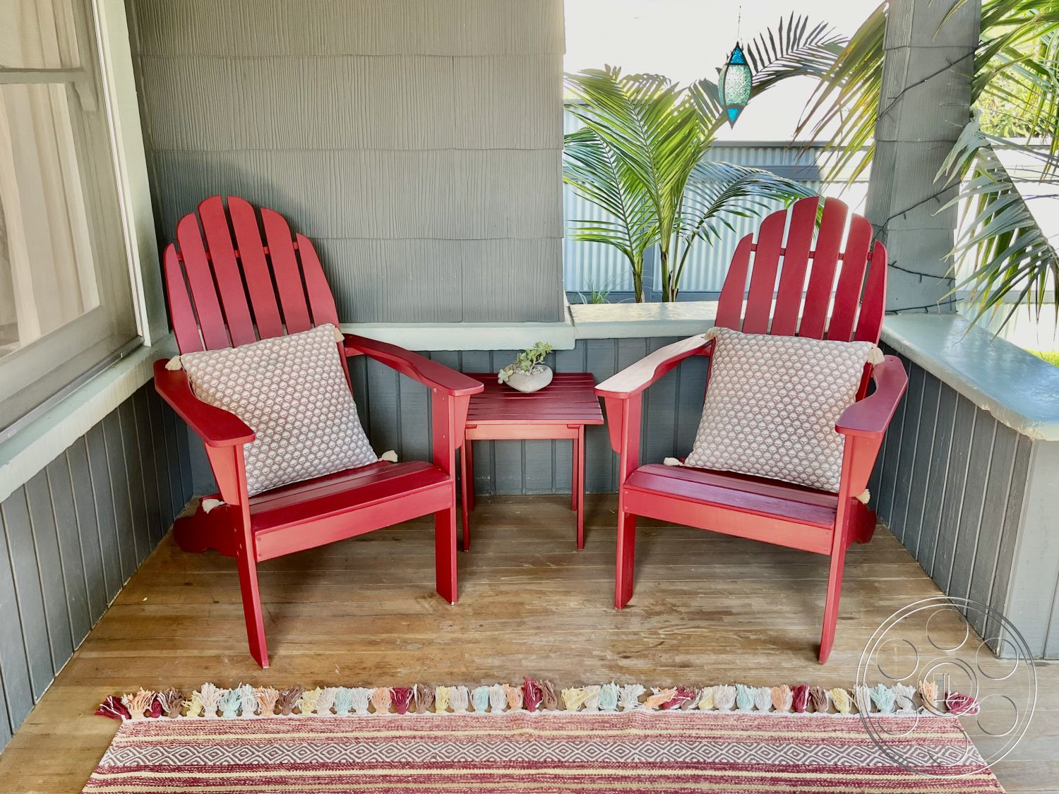 Craftsman 56 - exterior, casual seating area, light wood decking, textured wood paneling, small wooden side table, natural light outdoor space, red wooden adirondack chairs, tan accent pillows, outdoor area rug, green palm plants, cozy porch setting, decorative lantern pendant, comfortable outdoor furniture