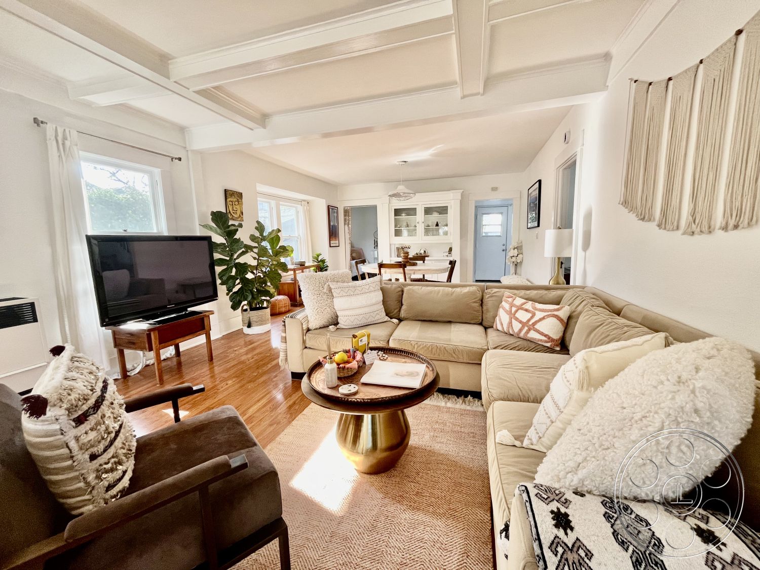 Craftsman 56 - neutral color palette, hardwood flooring, coffered ceiling, round coffee table, textured area rug, open floor plan, natural light from large windows, decorative throw pillows, beige sectional sofa, dining area in background, interior living room, indoor potted plant, macrame wall hanging, TV on wood stand, urban home decor