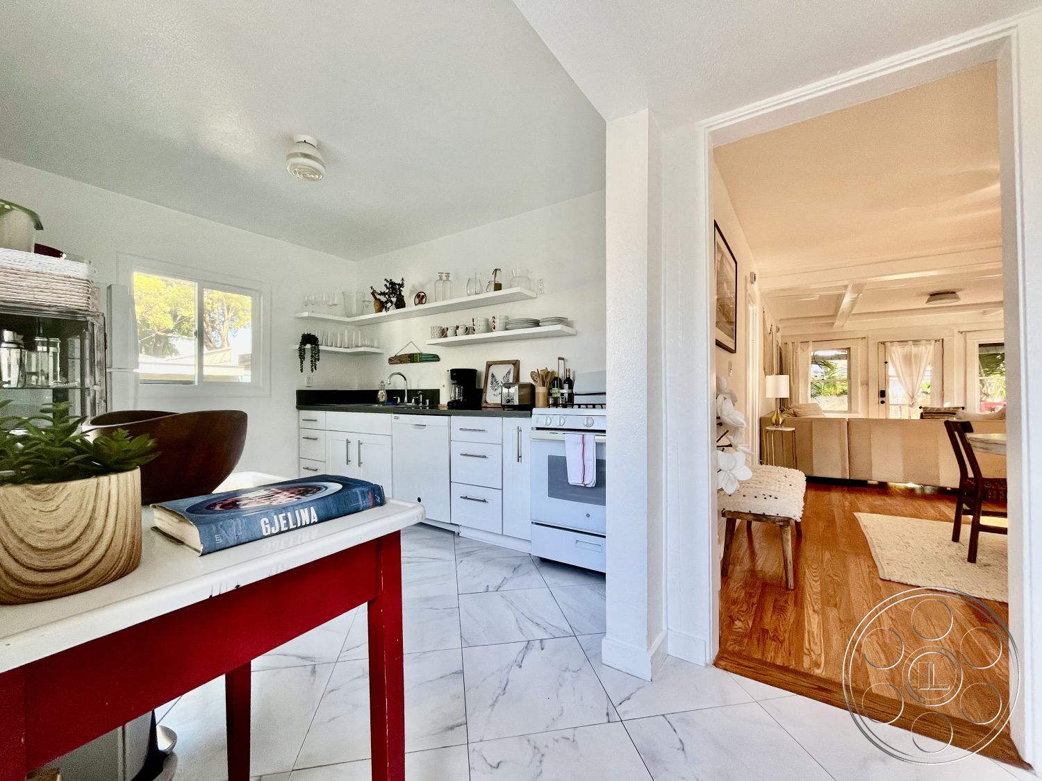 Craftsman 56 - black granite kitchen countertops, stainless steel appliances in kitchen, interior kitchen with marble tile flooring, white painted walls in kitchen, modern cabinetry with sleek handles, open shelving with glass dishware, natural light from window providing a view, red wooden kitchen table with circular legs, hardwood flooring in adjacent living area, cozy seating area with plush beige couch, bright lighting fixtures providing ambience, minimalist kitchen decor with potted plants, large open doorway leading to living room, stylish kitchen layout promoting flow, contemporary design elements across the space