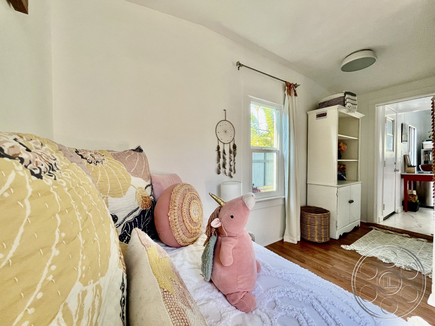 Craftsman 56 - contemporary decor, textured bedding, woven storage basket, wooden shelving unit, natural light from window, window with sheer curtains, hardwood bedroom floors, white painted walls, pastel decorative pillows, wall-mounted dreamcatcher, light fixture ceiling mount, doorway to adjacent room, cozy bedroom nook, interior guest bedroom