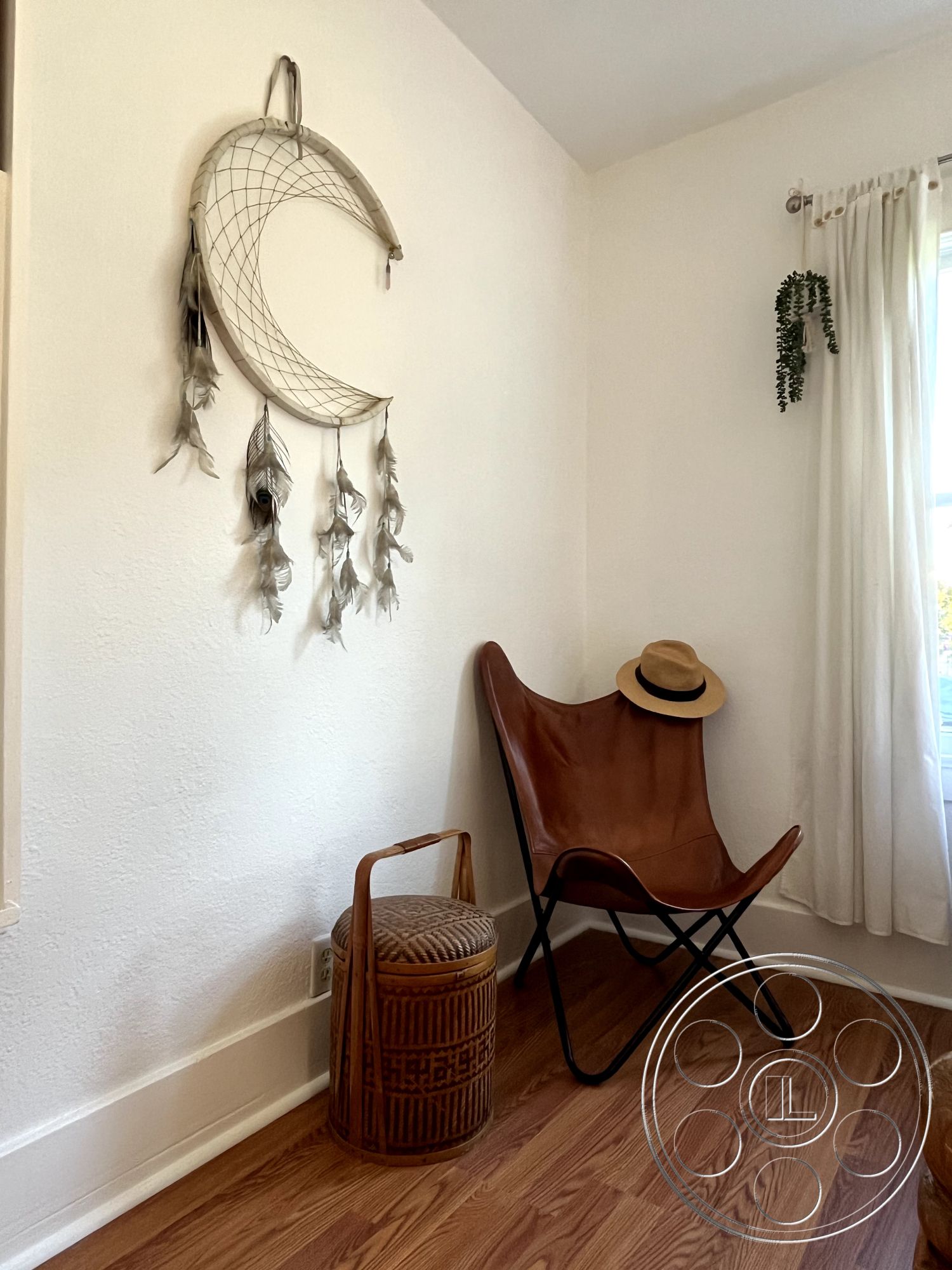 Craftsman 56 - natural light from window, window with sheer curtains, hardwood living room floors, minimalist decor style, white painted walls, feather wall hanging decor, butterfly chair with leather upholstery, woven rattan side table, decorative hat on chair, indoor plant in corner