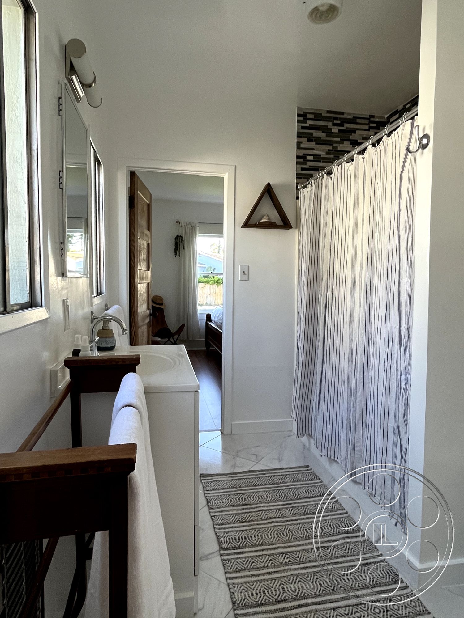 Craftsman 56 - framed mirror, natural light from window, glass shower doors, interior bathroom, white painted walls, textured tile shower wall, marble bathroom flooring, decorative patterned rug, wooden bathroom vanity, metal towel bar, striped shower curtain, doorway to bedroom, triangle wall decor