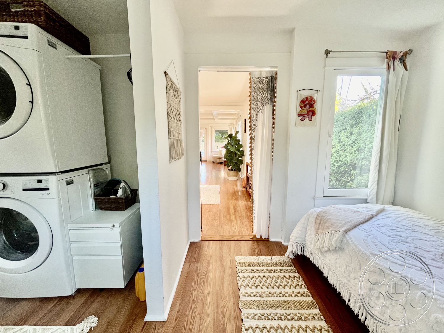 Craftsman 56 - natural light from windows, window with sheer curtains, interior laundry room with white appliances, hardwood flooring in hallway, white painted walls in hallway, textured wall hanging art, open doorway leading to living space, built-in storage above washer dryer, woven decorative throw on bench, green indoor plant for decor, striped area rug in hallway