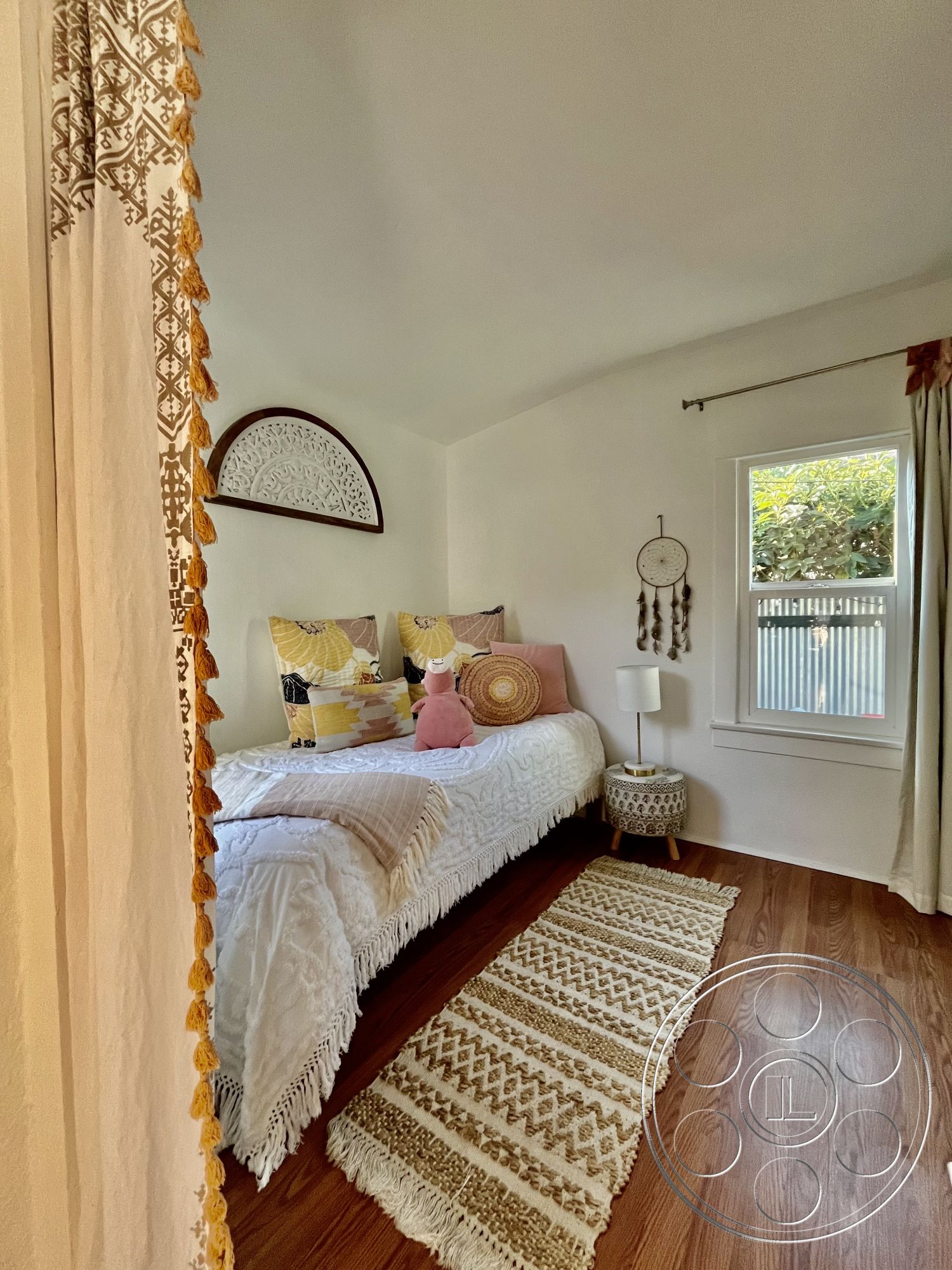 Craftsman 56 - natural light, hardwood flooring, woven area rug, colorful throw pillows, cozy reading nook, interior bedroom, textured white walls, decorative fringed bedspread, bohemian wall hanging, simple nightstand lamp, wooden window trim, plush toy accent, dreamcatcher wall decor, tropical-inspired decor, modern boho style