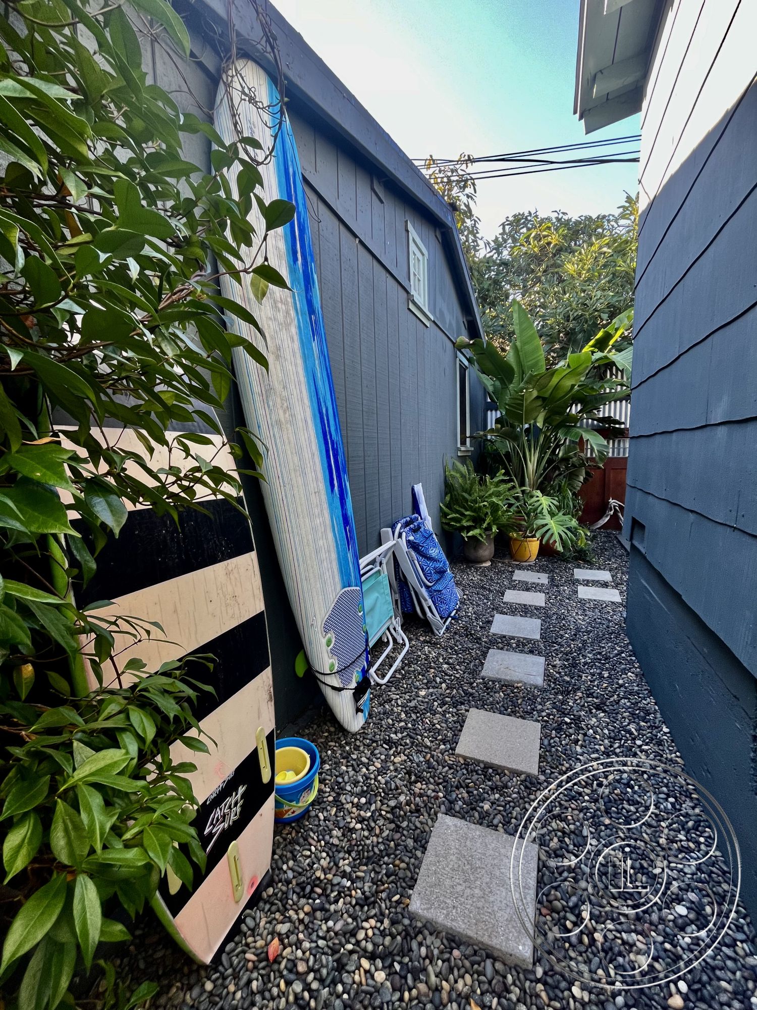 Craftsman 56 - natural light, exterior, stone stepping stones, casual outdoor setting, fenced backyard, blue painted wood siding, loose pebble pathway, tropical plants landscaping, surfboards against wall, beach chair storage, garden container pots, narrow side yard layout, coastal home environment