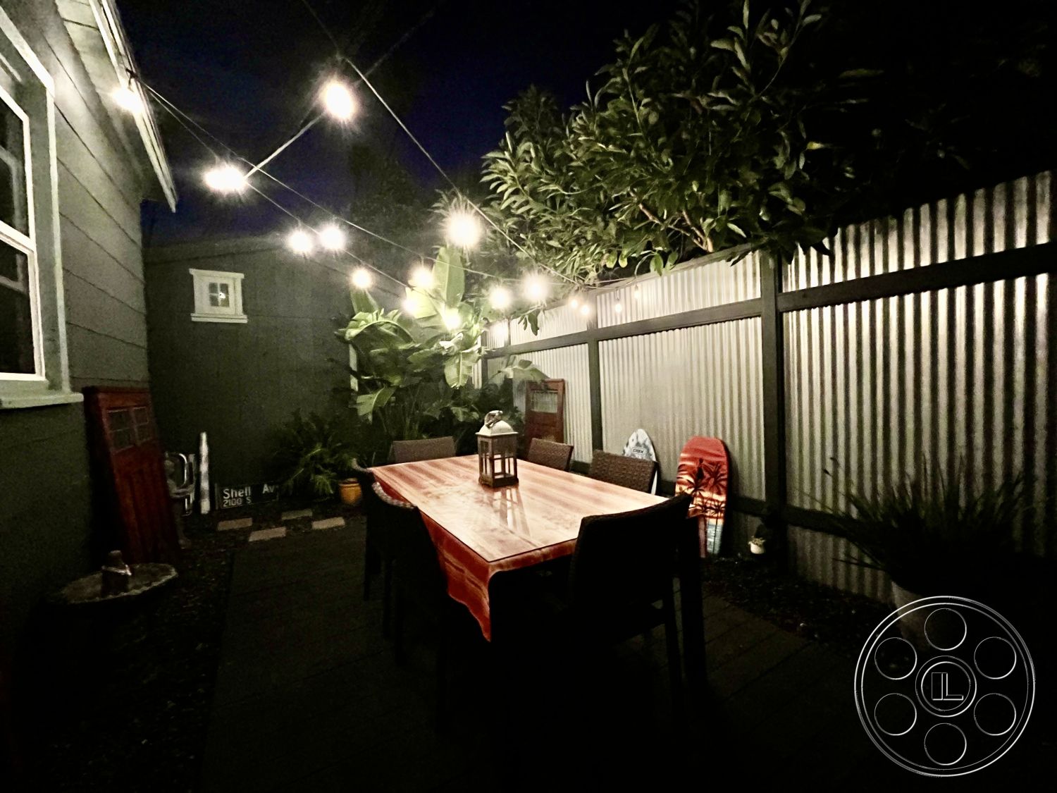 Craftsman 56 - wooden dining table, tropical landscaping, metal outdoor chairs, exterior patio area, textured wall details, potted plants decor, string lights overhead, modern outdoor dining space, corrugated metal fence, dark wood decking, lantern accent lighting, nighttime outdoor setting