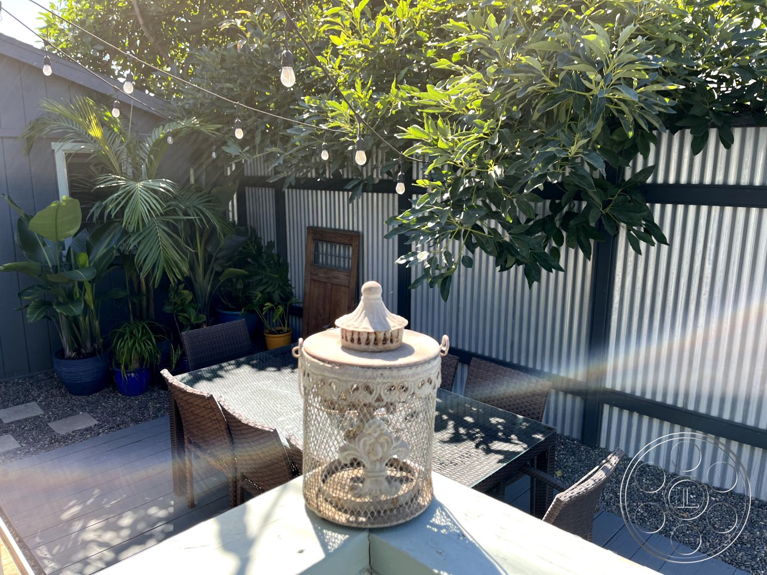 Craftsman 56 - modern farmhouse style, gravel ground cover, green foliage backdrop, landscaped garden area, exterior patio area, wood deck flooring, rectangular outdoor dining table, string lights overhead, woven wicker dining chairs, decorative metal lantern, colored planters with tropical plants, corrugated metal privacy fence, suburban outdoor retreat, sunlit outdoor living space