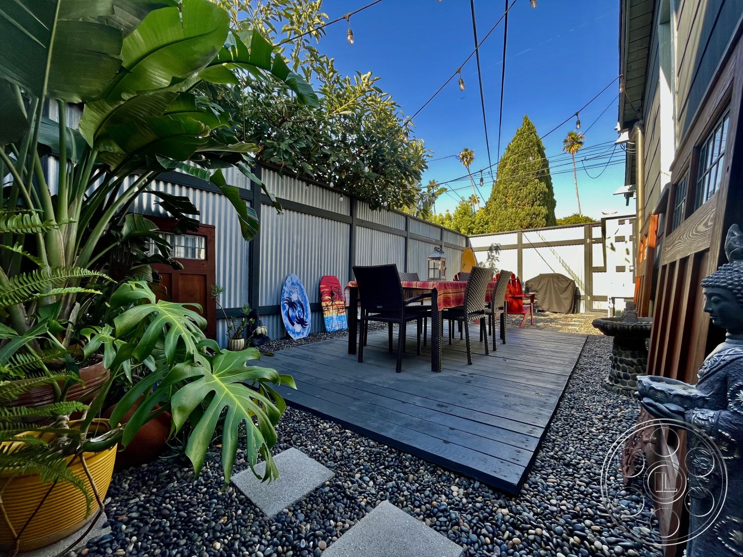 Craftsman 56 - suburban backyard, hedge privacy screen, sunny outdoor setting, wood deck flooring, contemporary outdoor dining area, string lights overhead, pebble stone landscaping, exterior courtyard patio, tropical plant pots, metal garden fencing, decorative stone Buddha statue, colorful surfboard wall art, modern outdoor chairs, wooden door accent, outdoor grill area